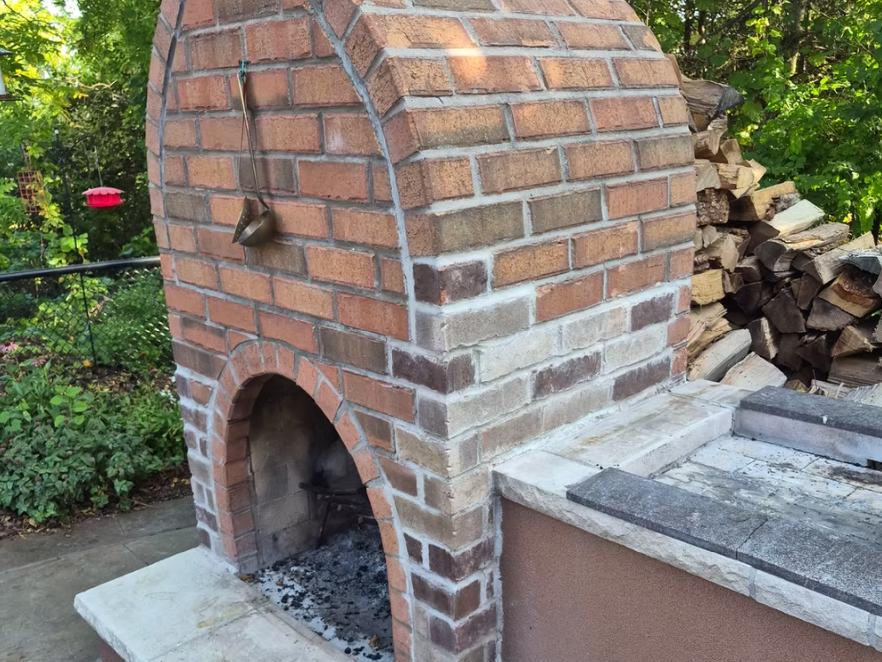 A brick outdoor fireplace with a small oven opening at the bottom, a chimney on top, and a wood pile beside it. There are stone steps in front and greenery in the background.