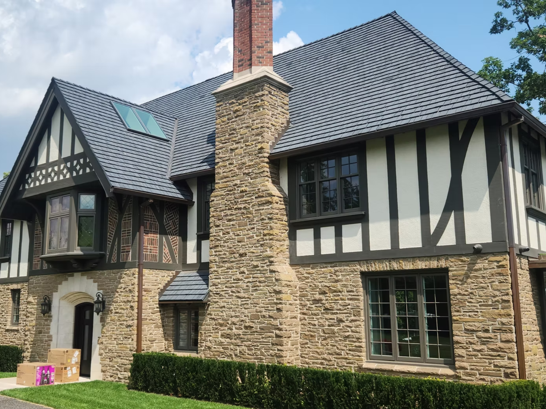 Front of a large house with stone and Tudor-style black and white woodwork, multiple windows, and a prominent brick and stone chimney under a partly cloudy sky.