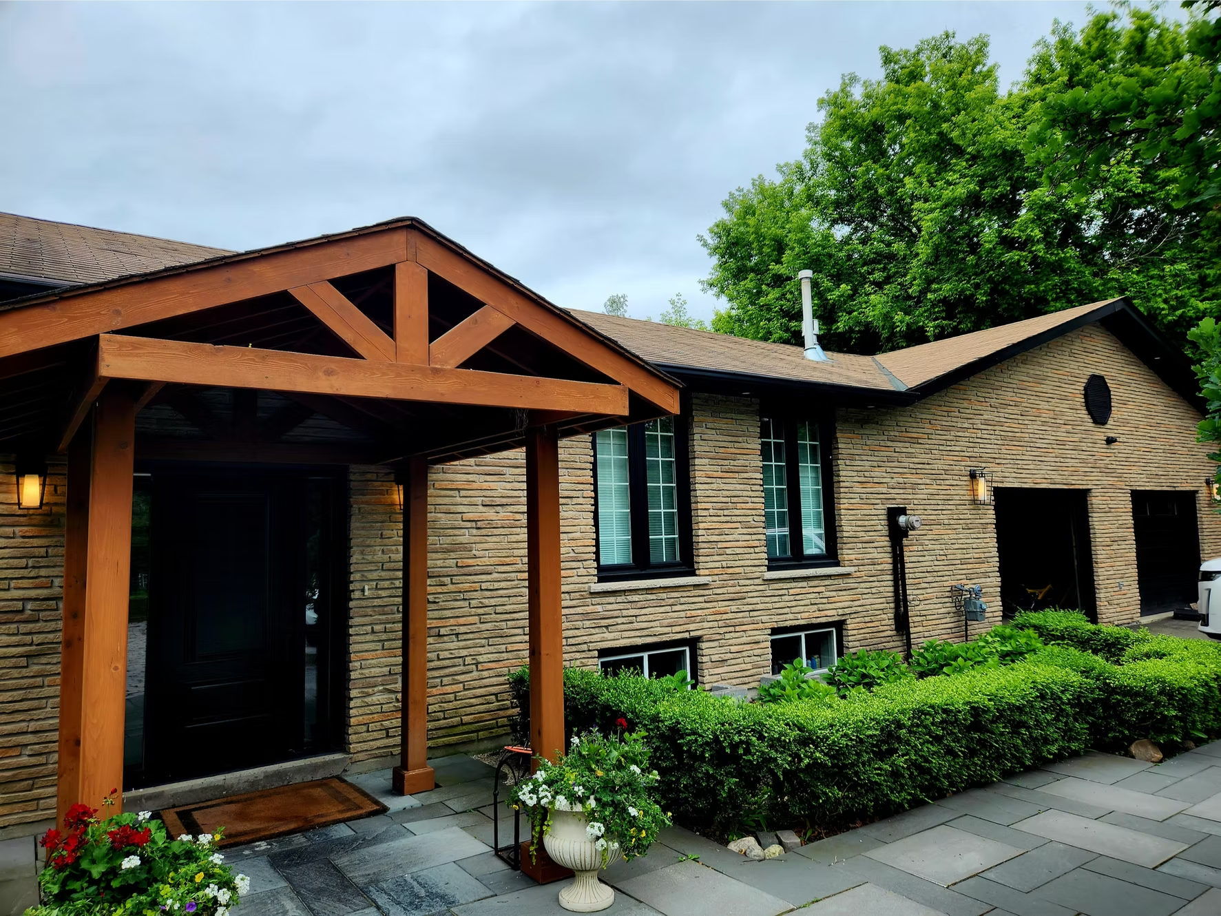 View of a brick house with a wooden trellis structure in the front yard, surrounded by lush green bushes and flowering plants, with large trees in the background and a cloudy sky overhead.