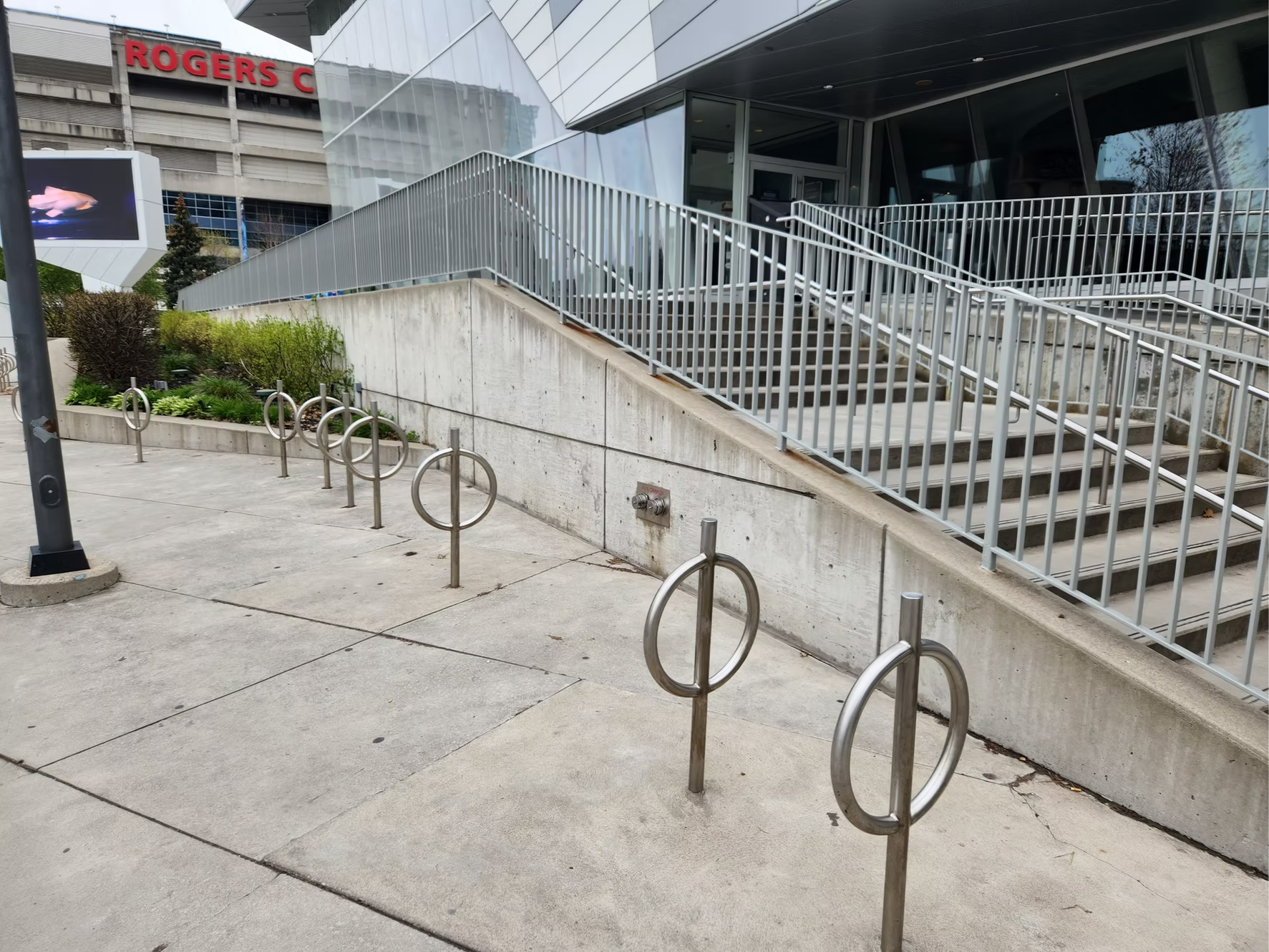 Stainless steel bike racks in front of a modern building with glass and metal exterior, with stairs and railing leading up to an entrance, and a large digital screen on the sidewalk.