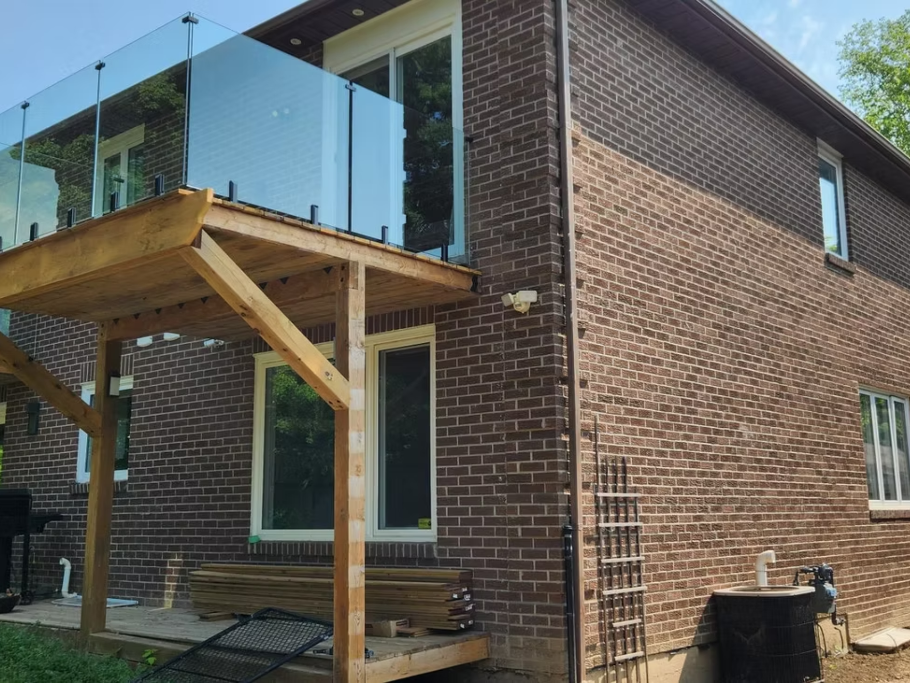 Backyard with an unfinished wooden deck extension, glass railing, brick house exterior, and construction materials underneath.