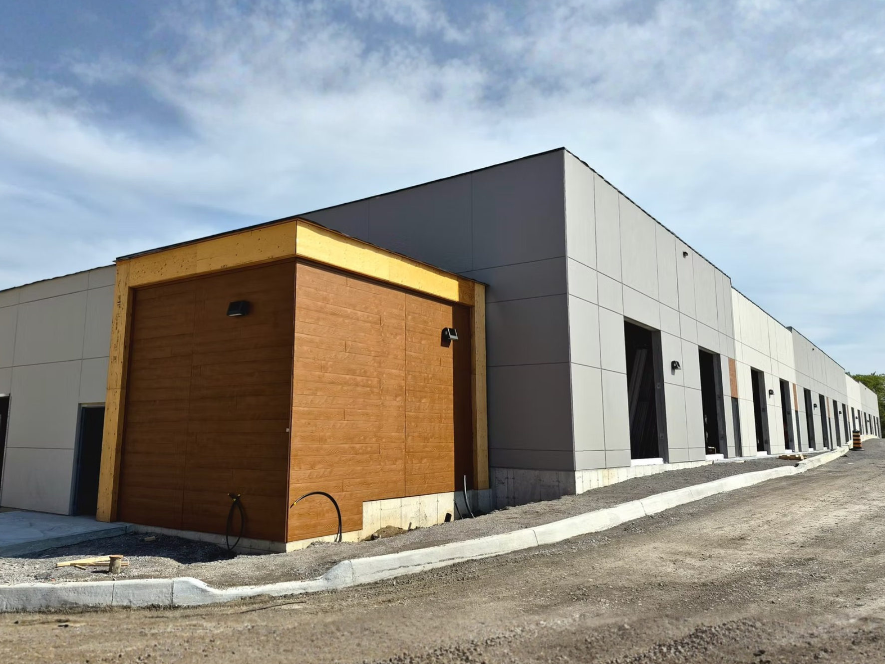 Modern building under construction with a combination of white and grey exterior panels, and a wooden section with a yellow trim near the entrance. The building has large windows and is situated along a dirt road.