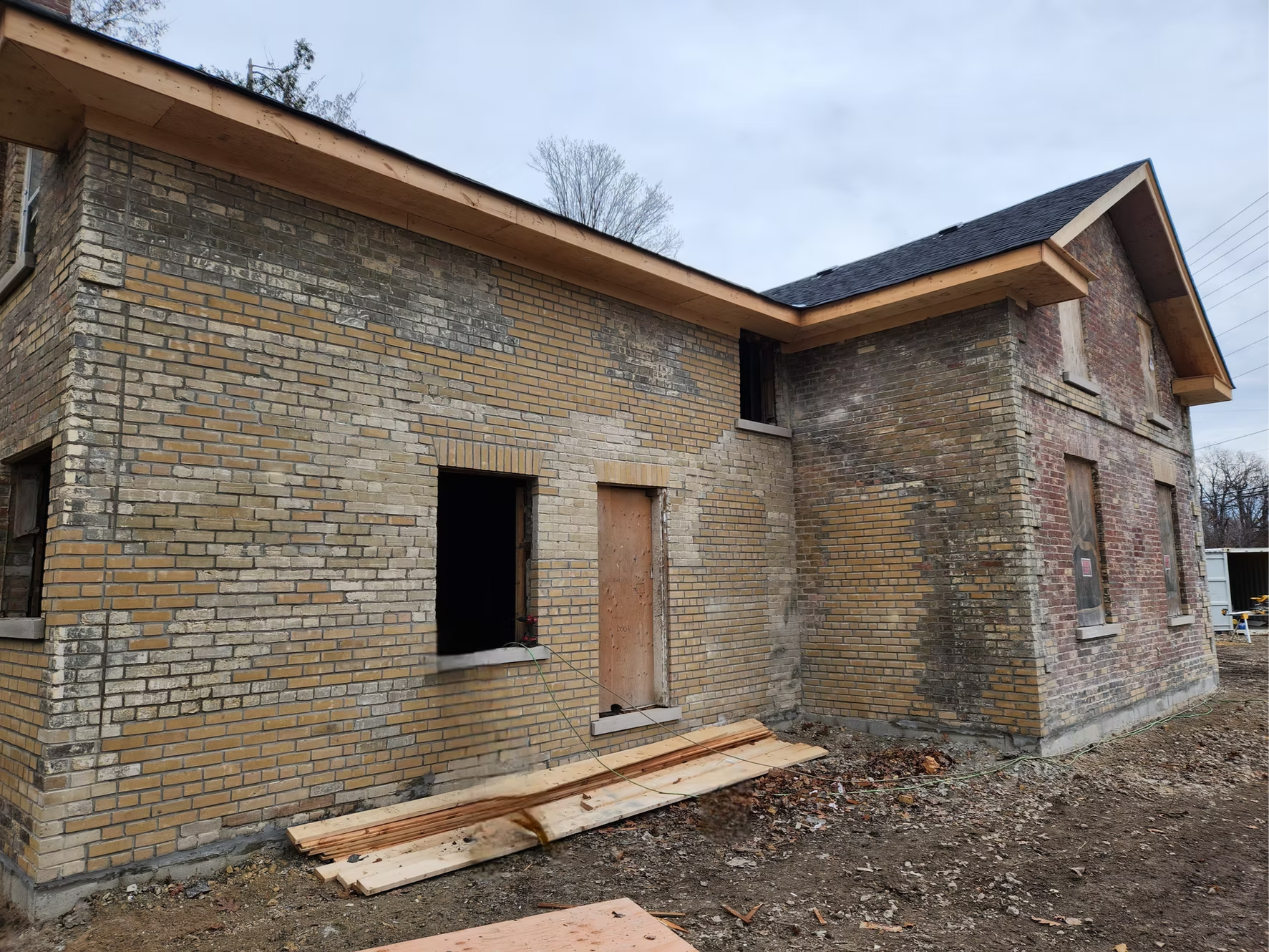 The image shows a brick house under construction with some sections of the brick exterior not yet completed. The house has a black shingle roof with exposed wooden eaves. There are two window openings and one door opening, all with unfinished frames. Construction materials and debris are scattered on the ground in front of the house.