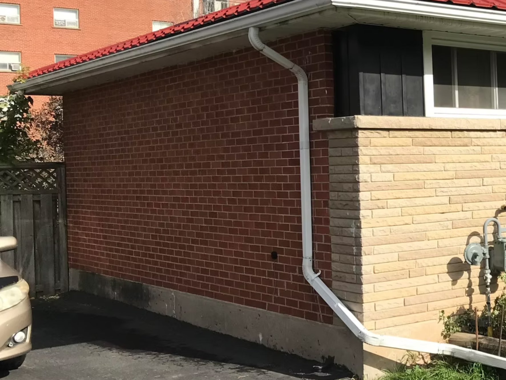 Part of a brick building with a beige brick section and a red brick section. A white drainage pipe runs down the corner. A window with a white frame is visible, along with an air conditioning unit in the background window. There is a small shrub and some utility meters on the ground, alongside a parking area.