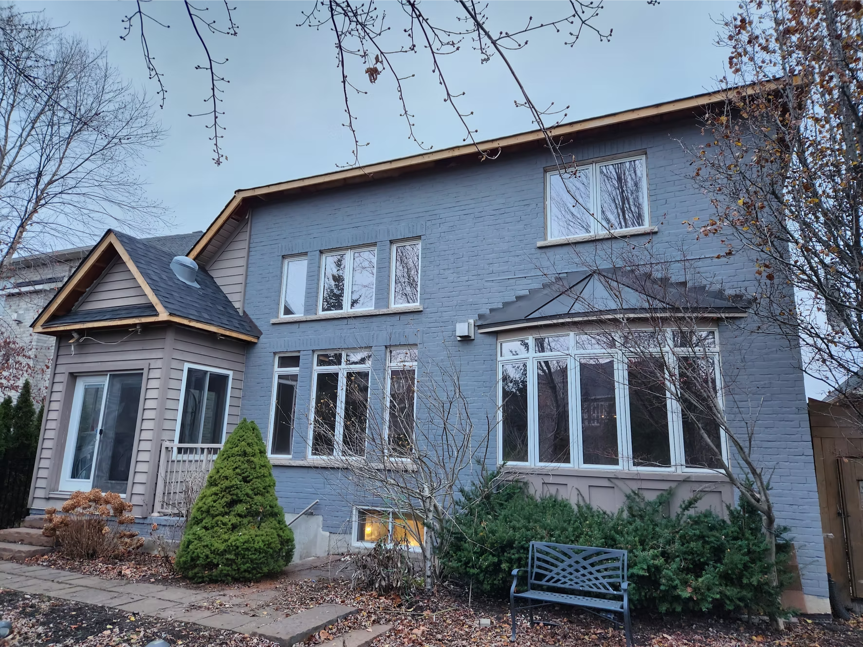 A large two-story house painted blue with a small attached porch area, surrounded by trees and bushes, during late fall or early winter.