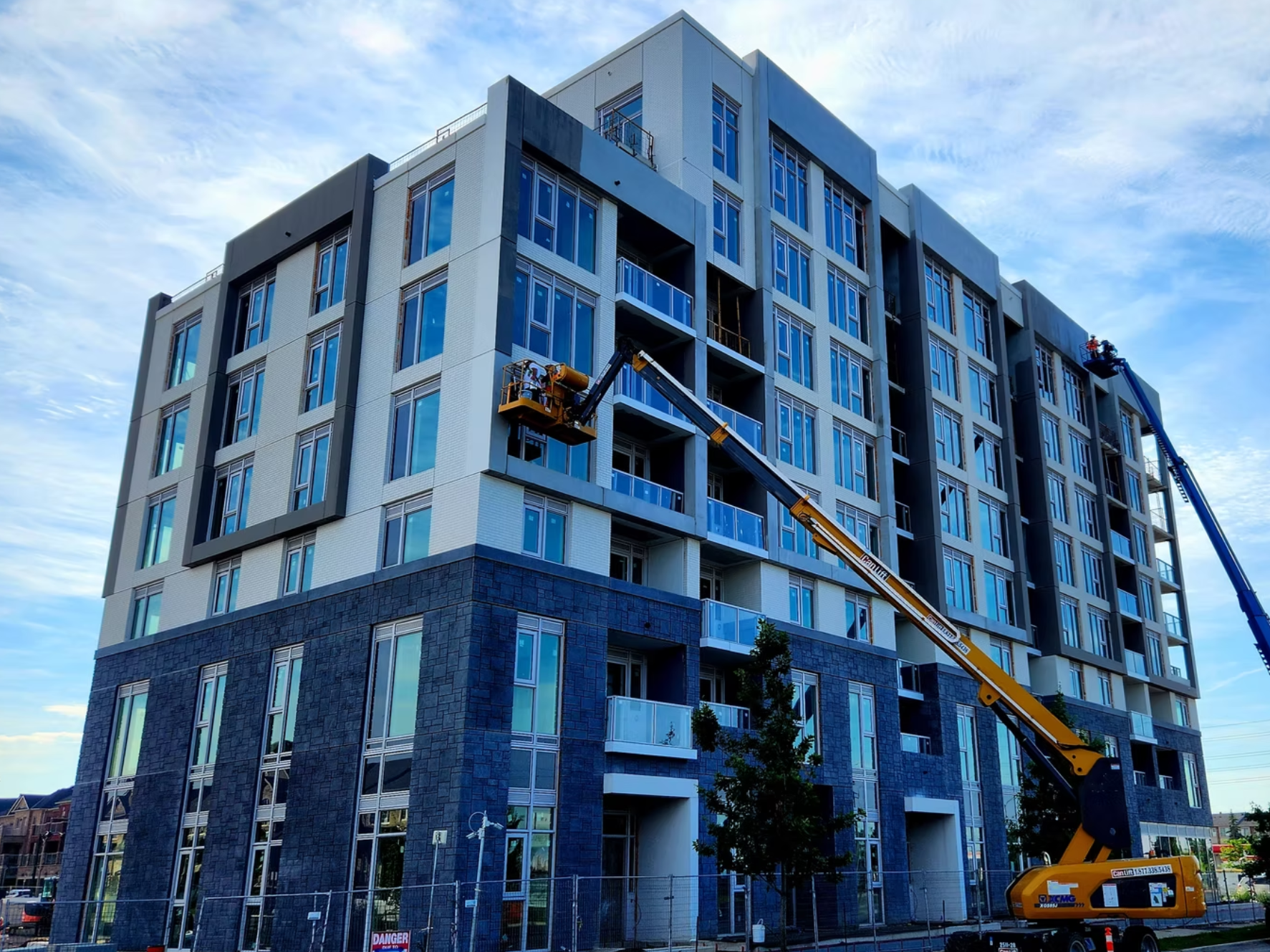 Construction workers on a cherry picker working on the exterior of a modern multi-story apartment building during daytime, with blue sky and clouds in the background.