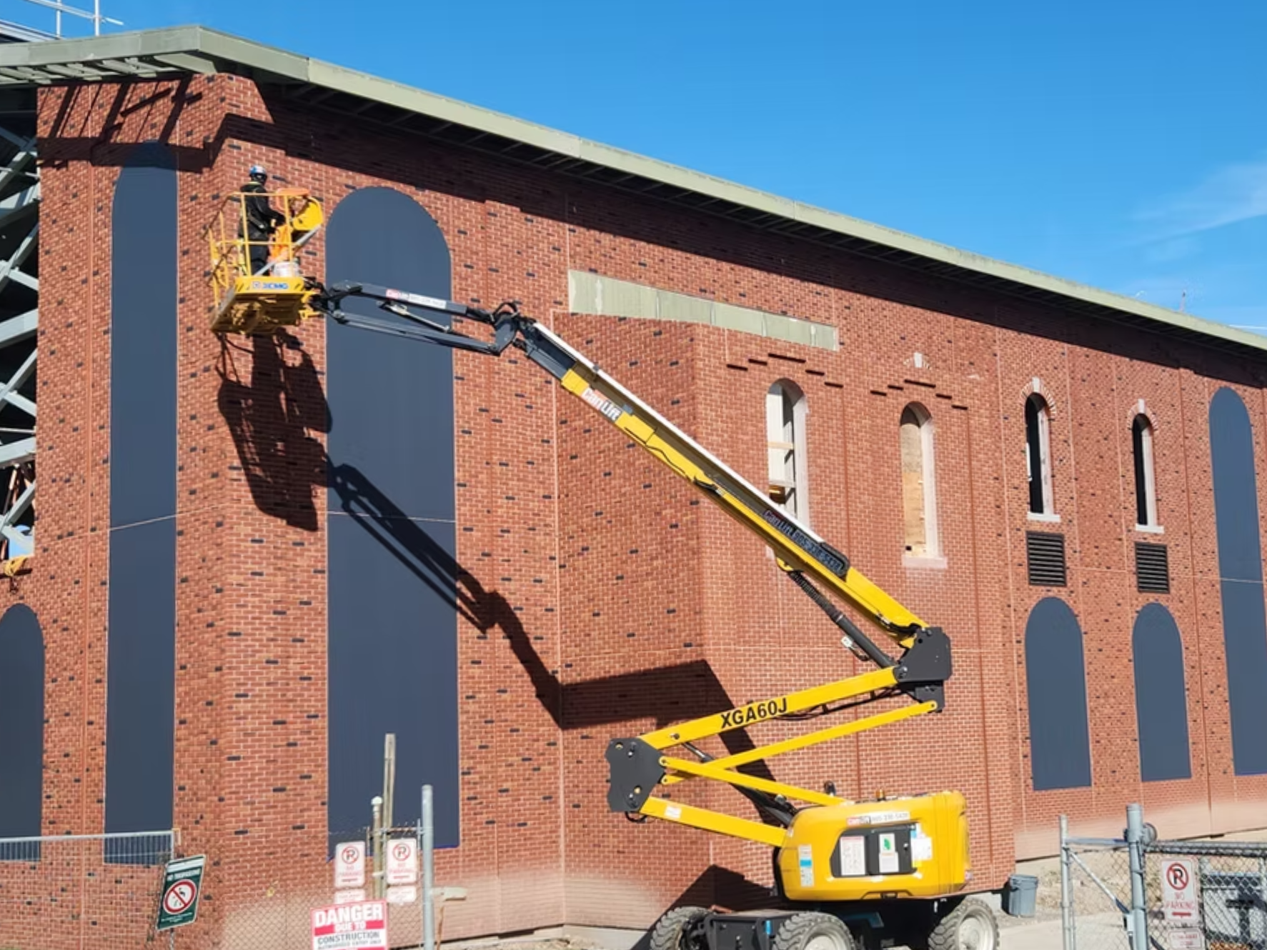 A worker on a yellow lift platform painting or repairing the upper part of a brick building under a clear blue sky.