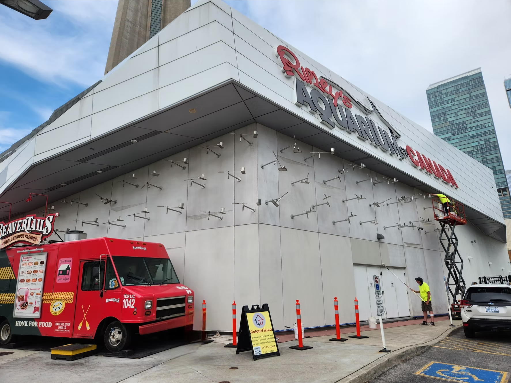 A large building with signs reading Ripley's Aquarium of Canada. There is a red food truck in front, a worker on a scissor lift, and another worker on the ground near orange traffic cones. The building has numerous metal fixtures on its exterior wall, and high-rise buildings are visible in the background.