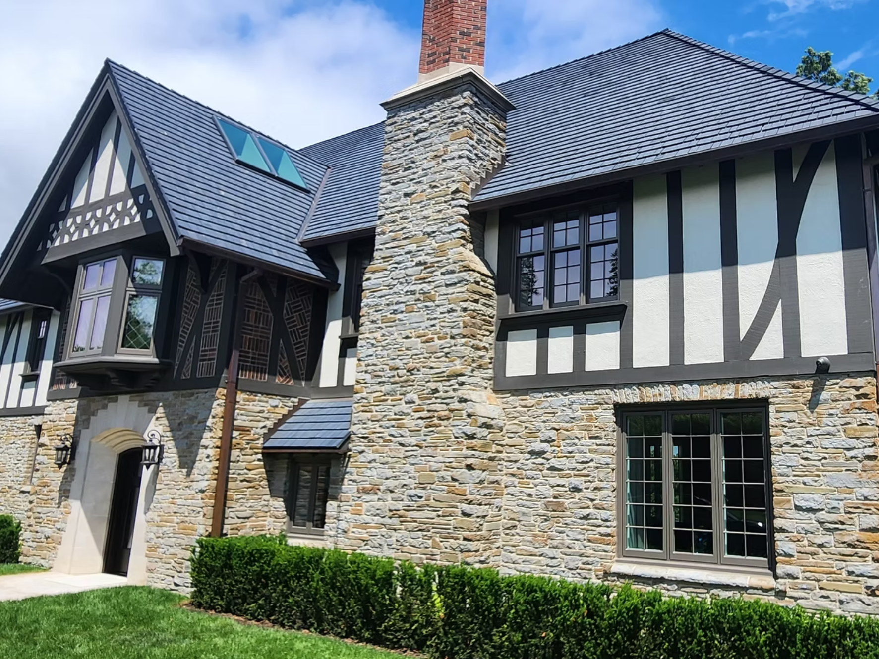 A large house with a stone and Tudor-style exterior, dark wooden beams, multiple windows, and a tall brick chimney, with a well-maintained lawn and neatly trimmed bushes in the front.
