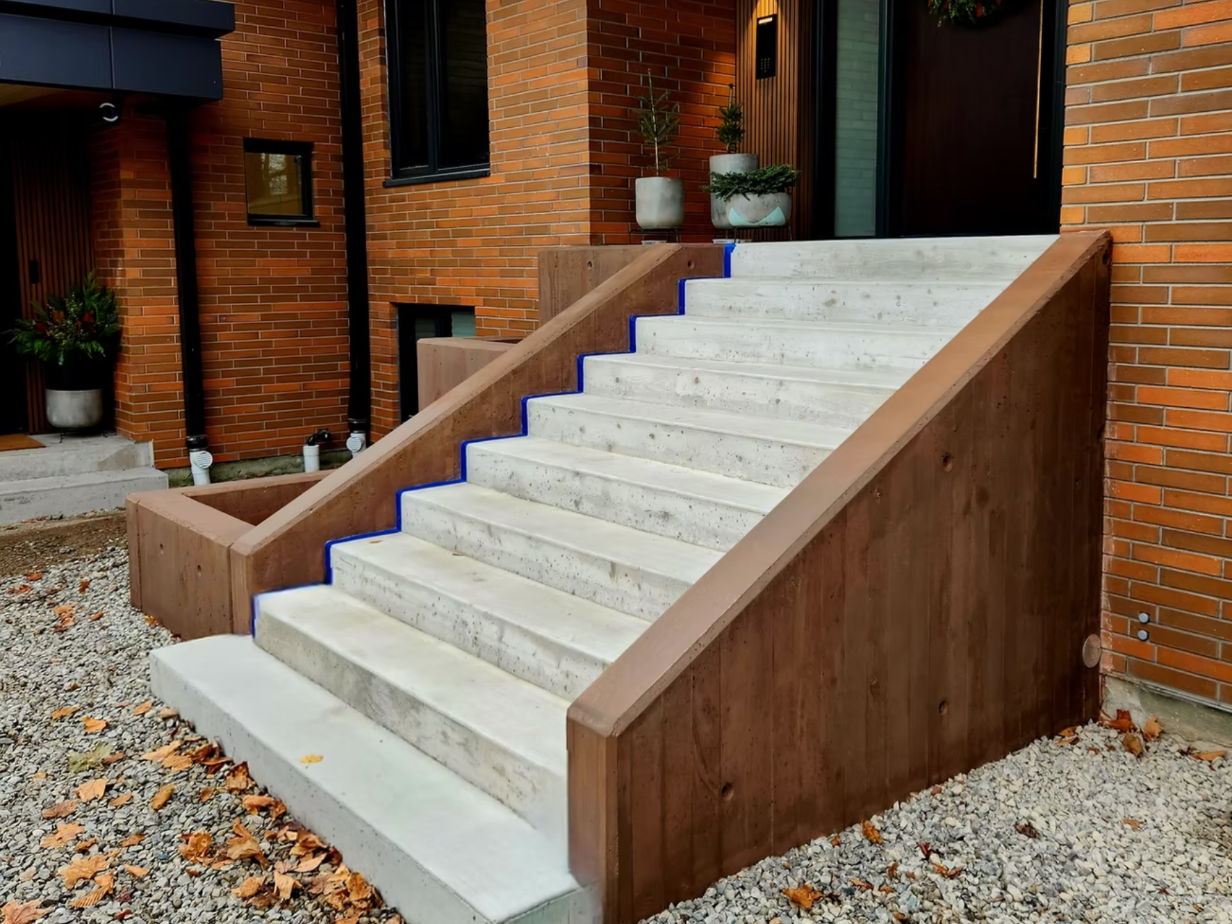 Concrete stairs with wooden sides leading up to a house entrance, decorated with potted plants on the landing. The house has red brick walls and black-framed windows and doors.