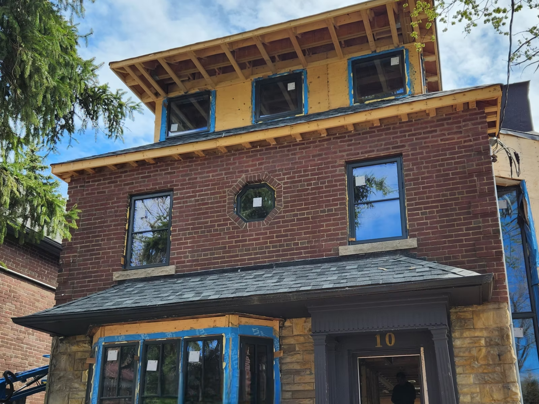 A multi-story house under construction with wooden framing, brick exterior walls on the lower levels, and installed windows. The house's upper level shows exposed wood and a new roof. The house number "10" is visible above the front door.