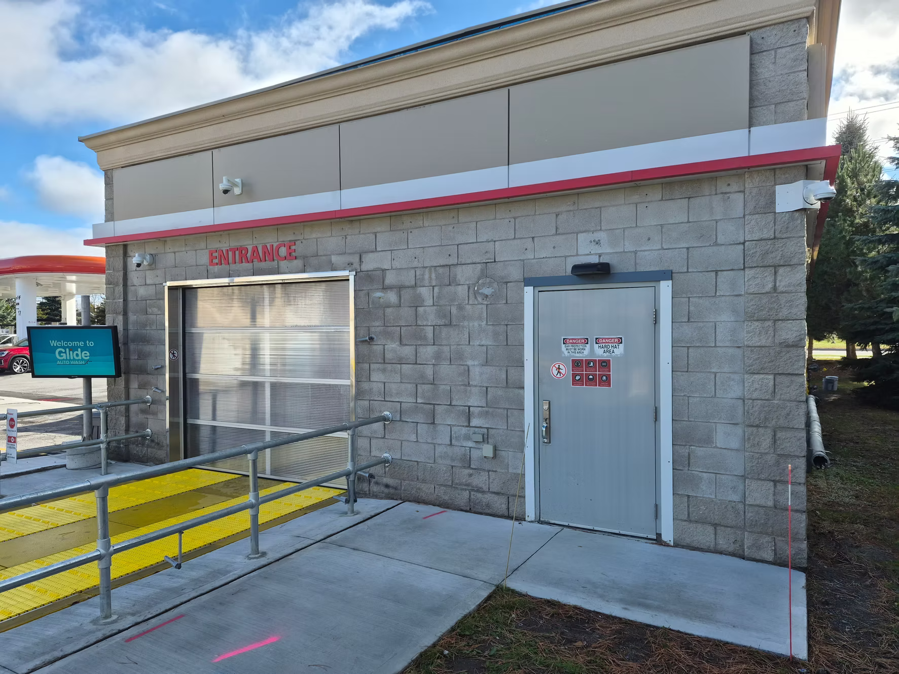 Exterior of a car wash facility with a large roll-up door, an entrance door with caution signs, security cameras, and a sign that says 'Welcome to Glide Auto Wash.' The area includes a wheelchair-accessible ramp with yellow tactile paving.