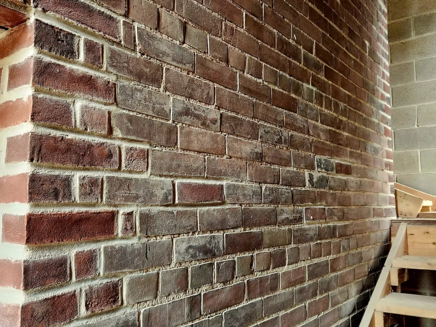 A brick wall with a mix of red and dark gray bricks, with some bricks showing signs of weathering or aging. Part of a wooden staircase is visible on the right side of the image.