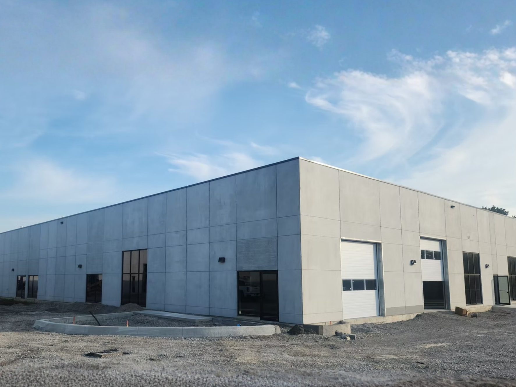 A modern commercial building under construction with a concrete exterior, large windows, and multiple garage doors, set against a blue sky with scattered clouds.