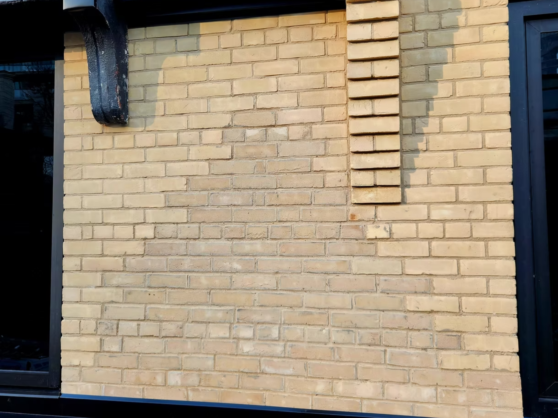 Close-up of a beige brick wall with a black metal drainpipe and a window with a black frame on the right side.