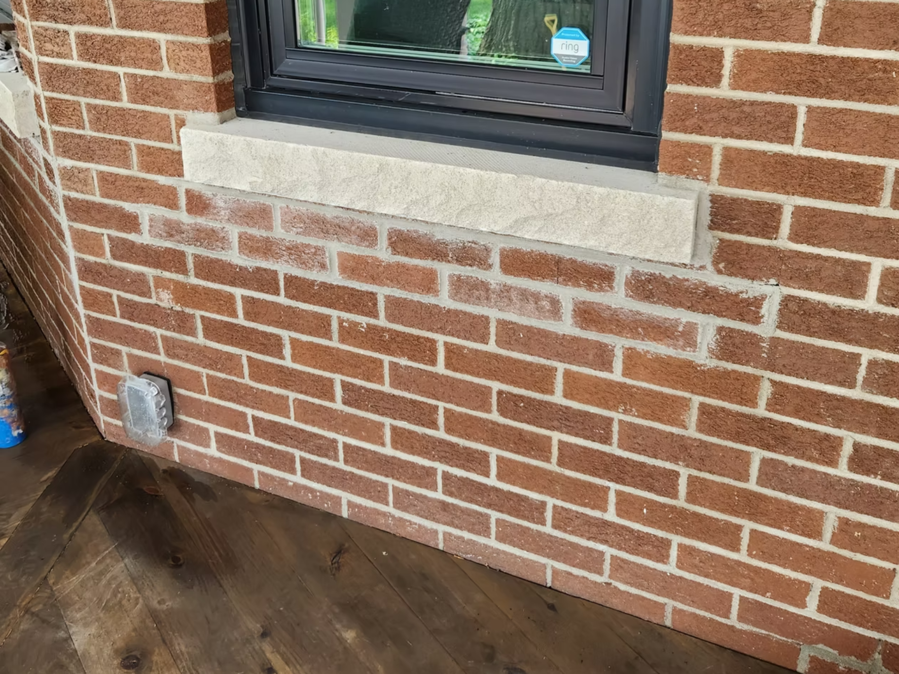 Close-up of a brick wall with a window featuring a black frame and a stone sill. On the left side, there's a corner of the wall with an electrical outlet installed near the baseboard. The floor is made of dark wooden planks.