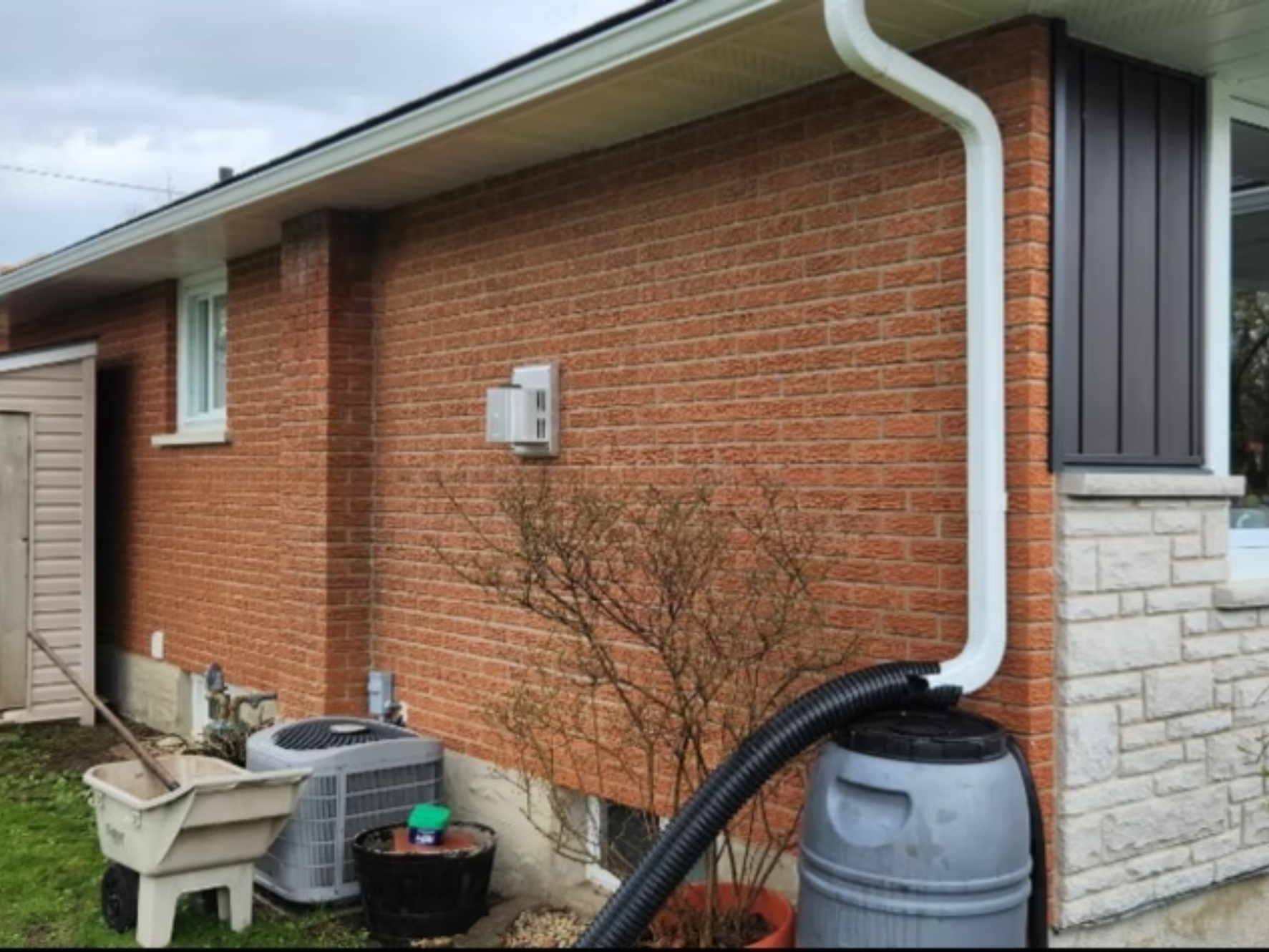Backyard of a brick house with a white sky and a gutter system. Items include a gray trash bin, black pots, a white plastic container, and a small leafless tree.