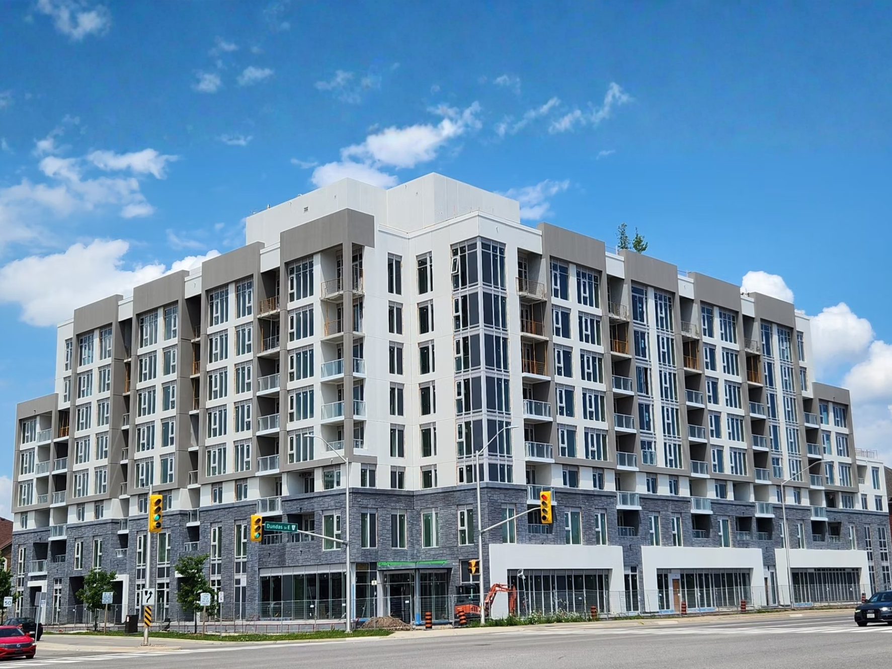 Modern multi-story residential building with glass windows and balconies, situated at an intersection with traffic lights, under a blue sky with some clouds.
