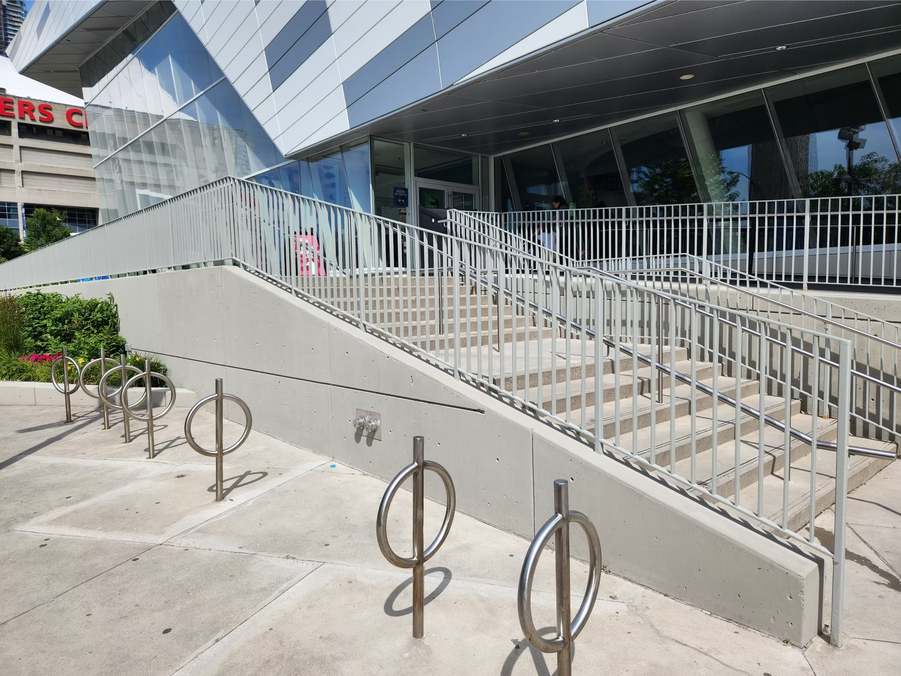 Modern building entrance with stairs, wheelchair ramps, and bike racks outside, with glass and metal architectural design.