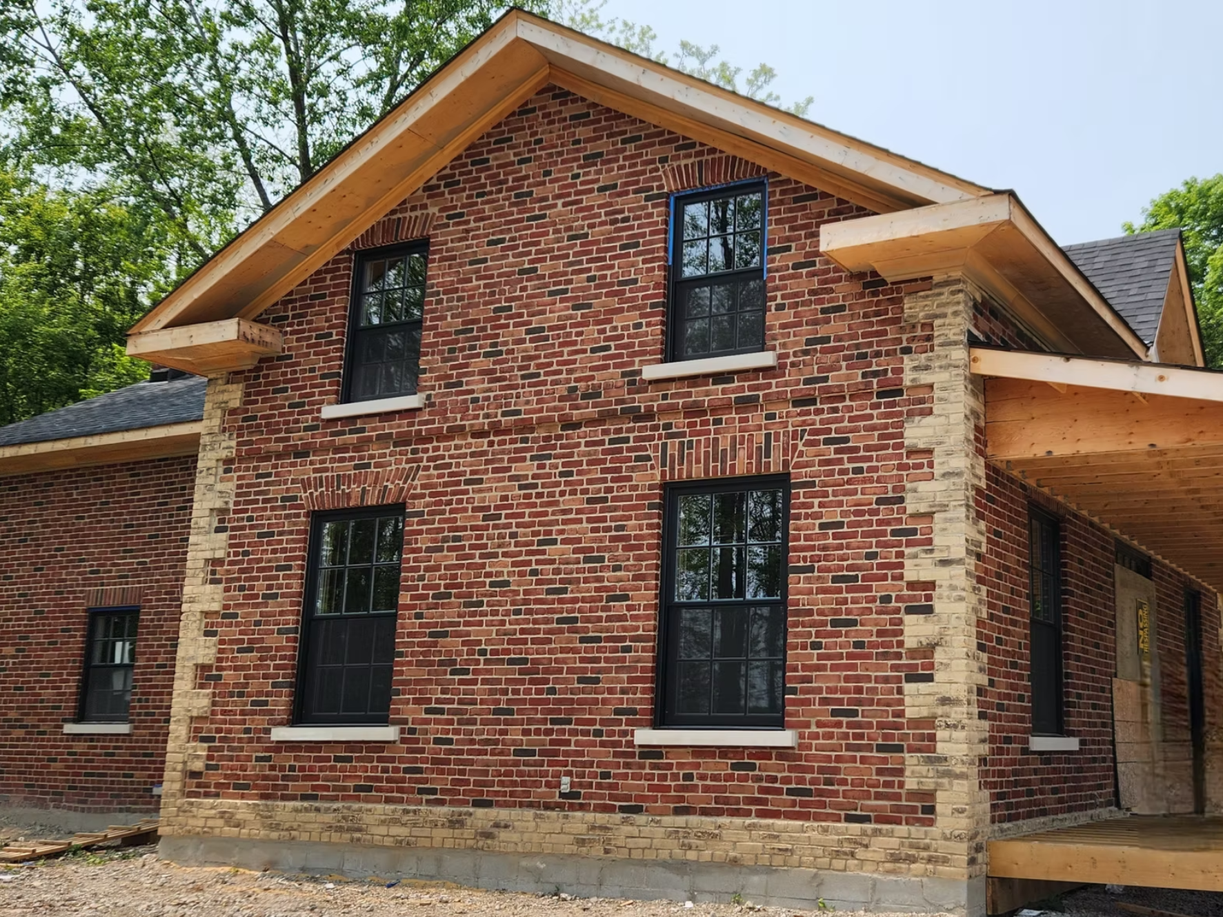 A brick house under construction with multiple black-framed windows, wooden trim, and a partially finished roof surrounded by green trees.