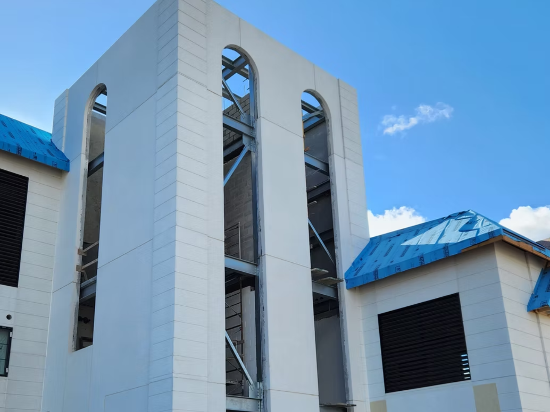 Residential building under construction with white exterior walls, black window covers, and blue roofing material, set against a blue sky with clouds.