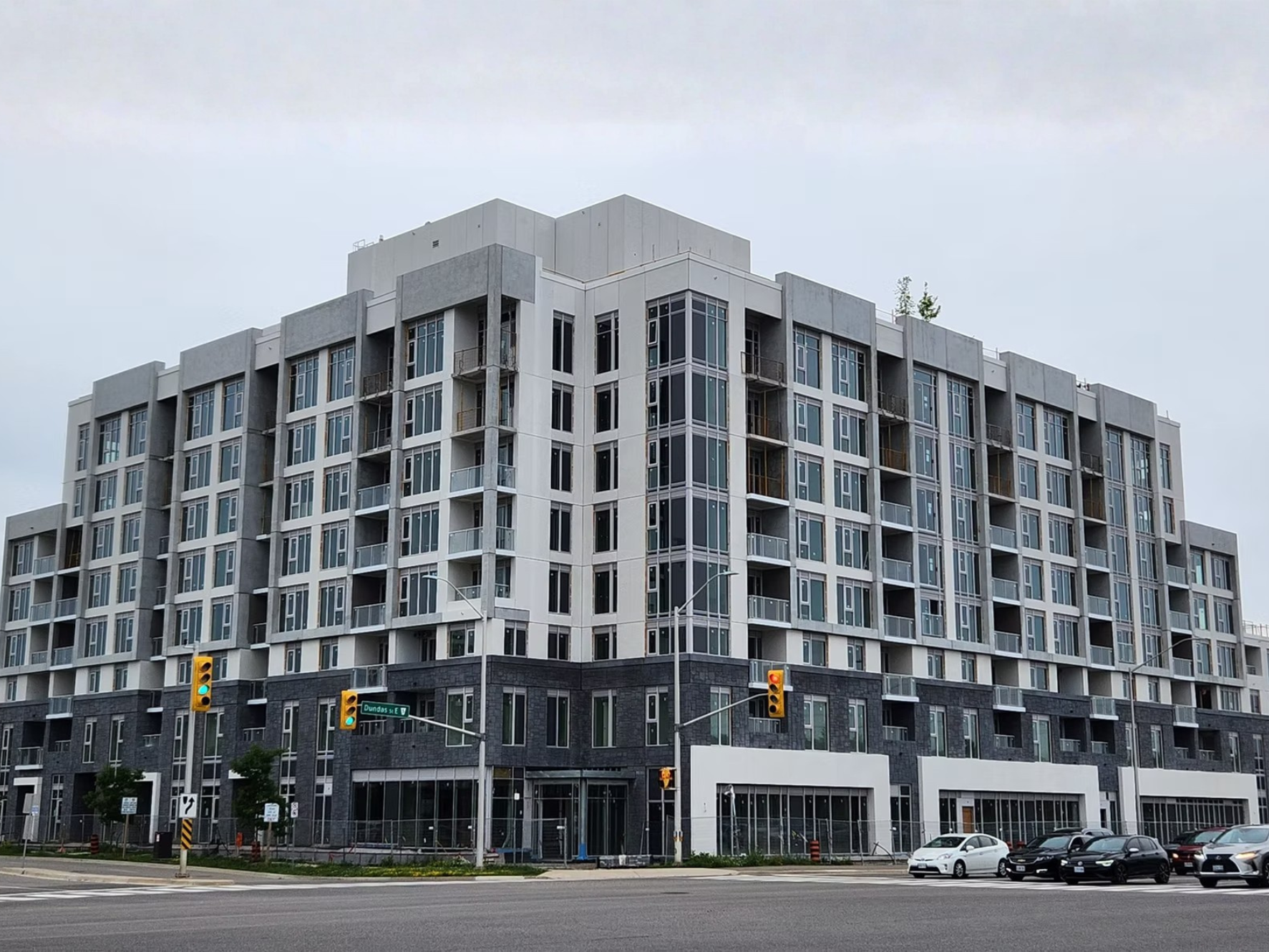 A tall modern apartment building with numerous balconies and large windows, located at a street corner with traffic lights, cars, and street signs.