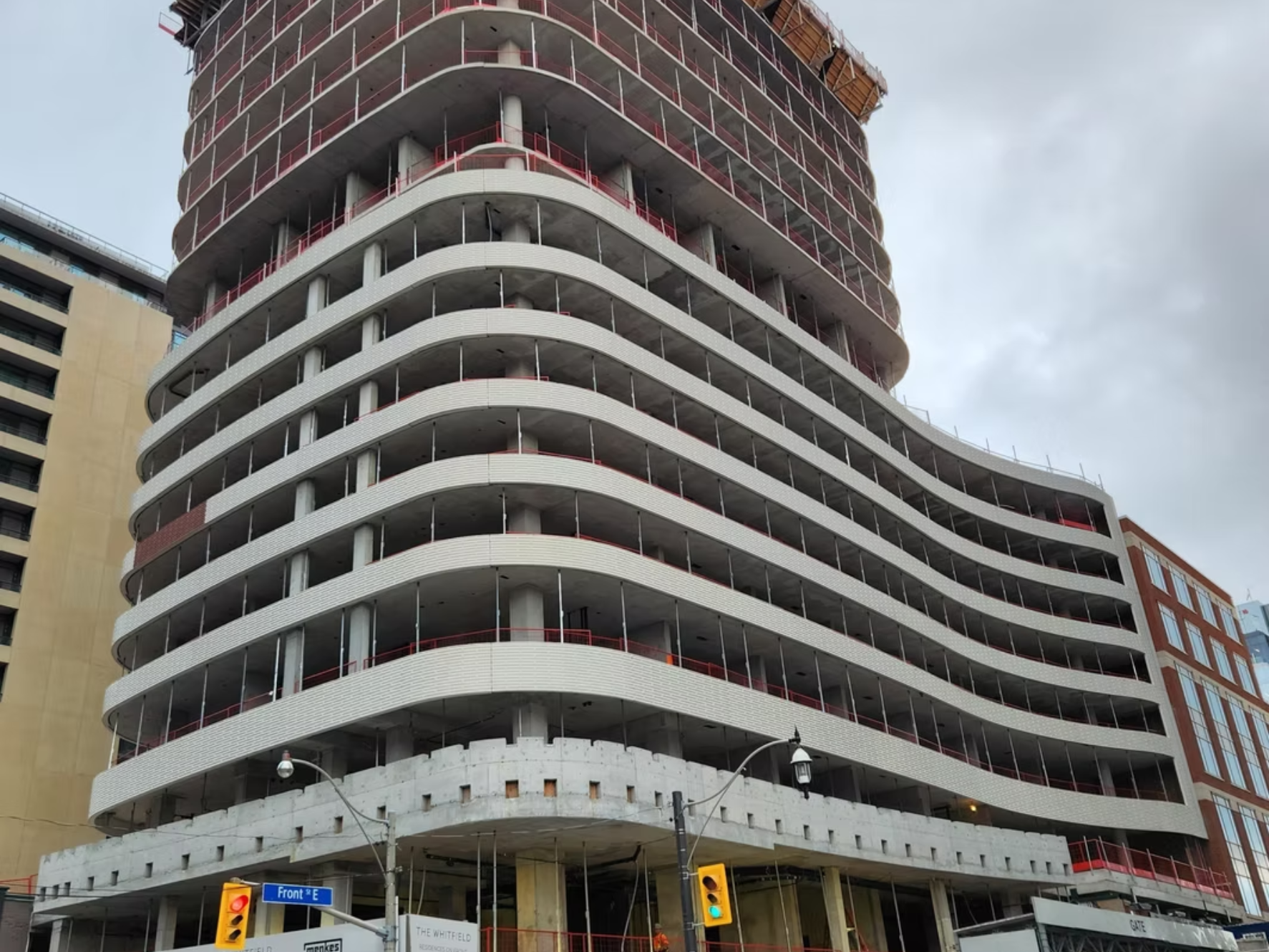 A multi-story building under construction with curved white balconies and red railings, surrounded by other completed buildings, with street signs and traffic lights in the foreground.