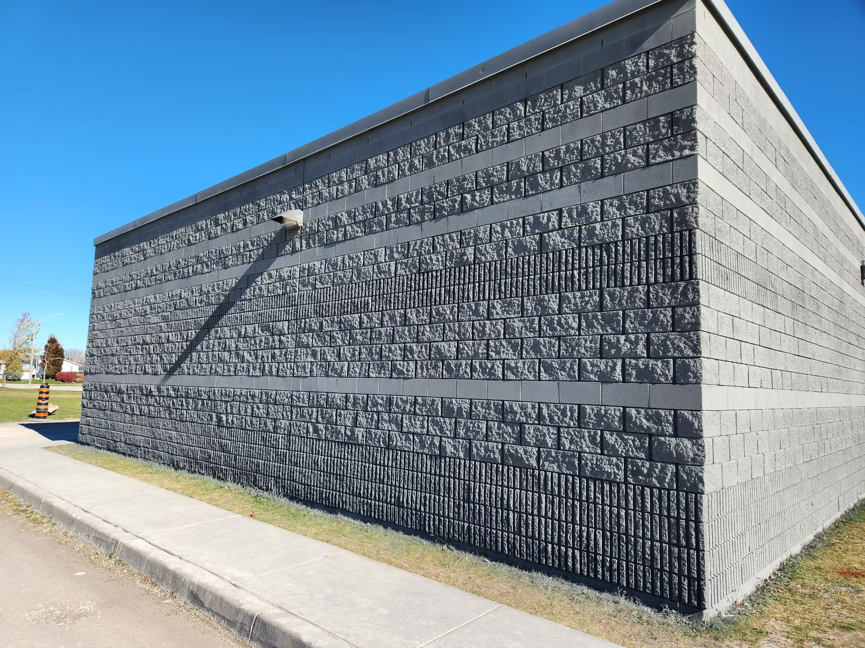 Exterior view of a large building with a brick wall facade, gray patterned bricks, and a security camera on the upper wall. Blue sky and a grassy sidewalk in the foreground.
