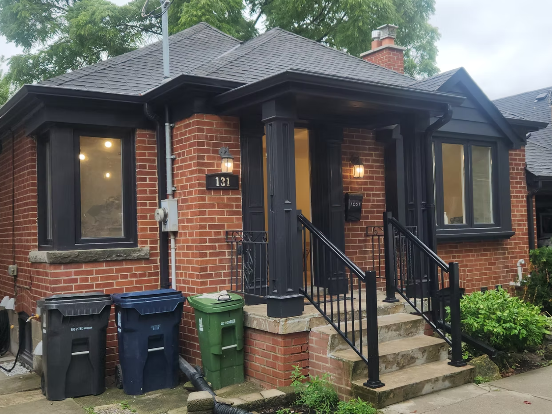 A brick house with a black front door and black window frames. The house number 131 is displayed on a black plaque next to the door. There are two outdoor wall lights on either side of the door. The front porch has black metal railings and concrete stairs. Several trash bins are placed at the bottom of the stairs. Green foliage and trees are visible around the house.