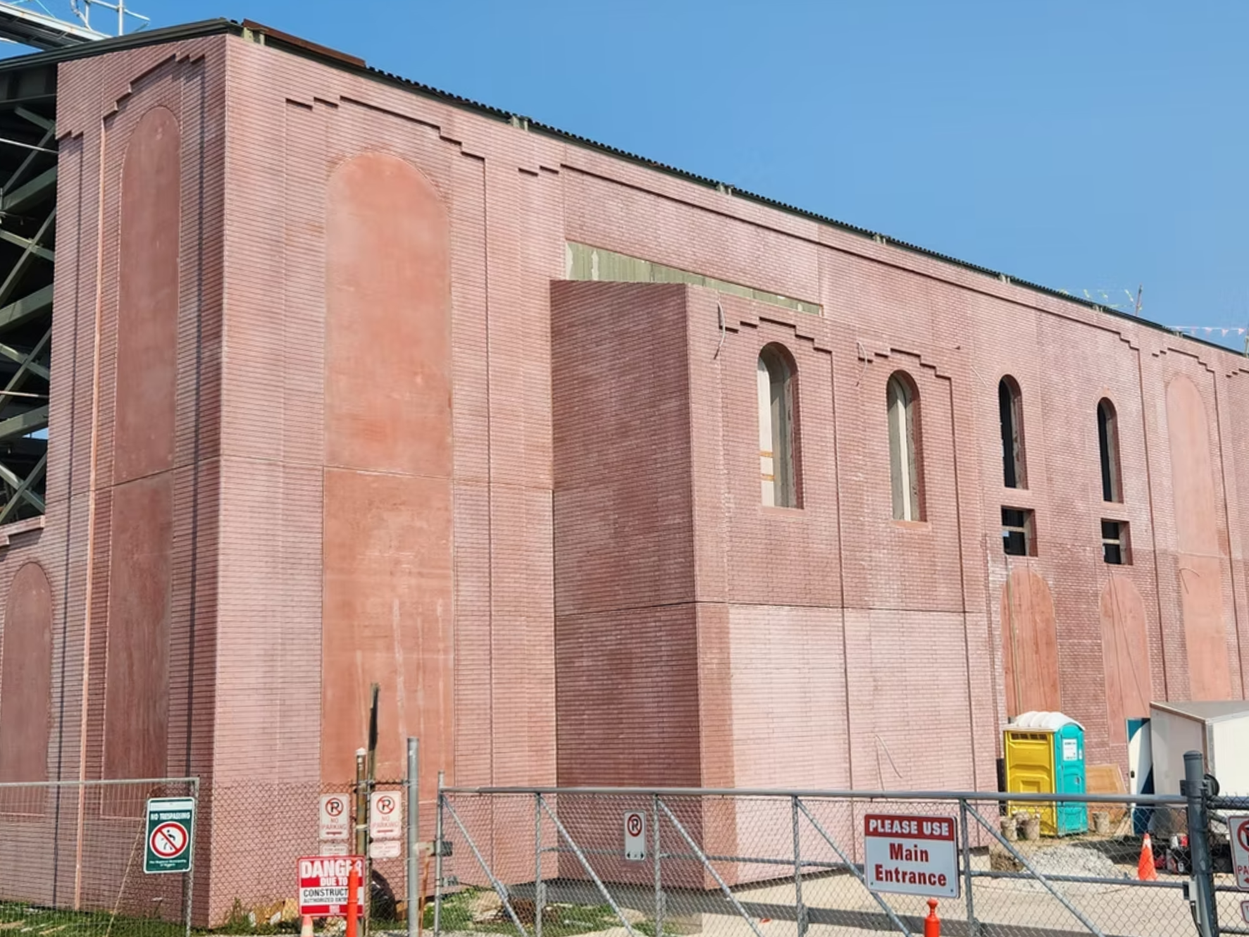 A large brick building under construction with a fenced perimeter and construction signs. The building features arched windows and decorative brickwork, with scaffolding visible on the side.