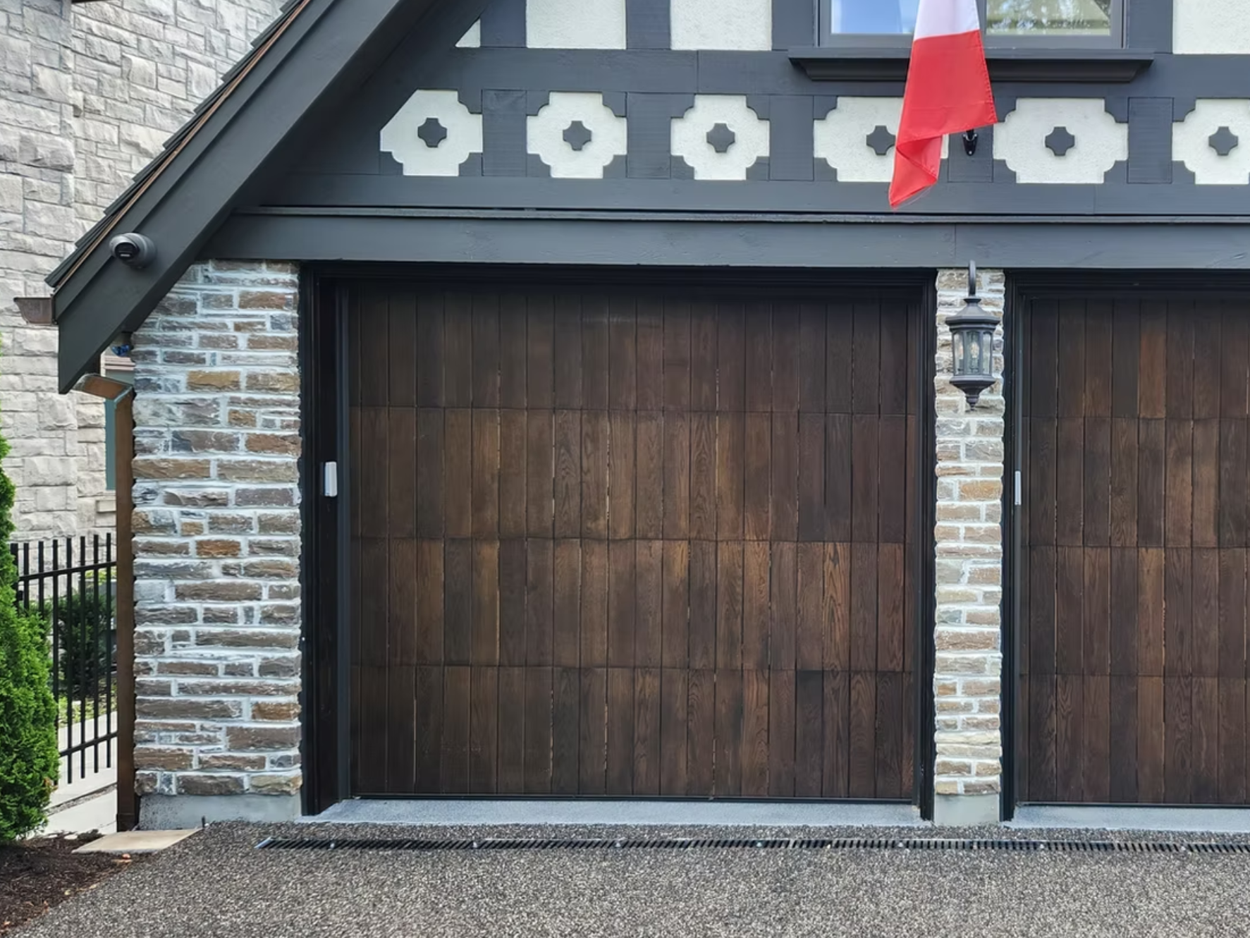Close-up of a garage door with wooden panels, flanked by brick columns, and a lantern-style light fixture on the right brick column, with a red and white flag hanging from the upper part of the right side.