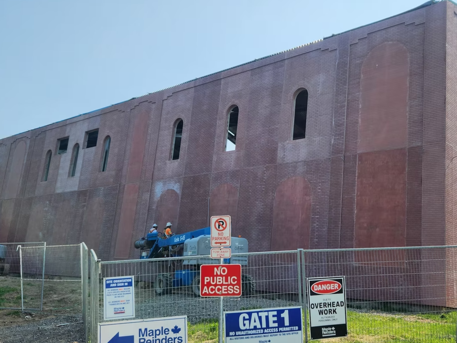 Construction site with a large brick building under renovation, surrounded by a metal fence with various warning and informational signs, including 'No Parking,' 'No Public Access,' and 'Gate 1.' Two workers are on a lift working on the building.