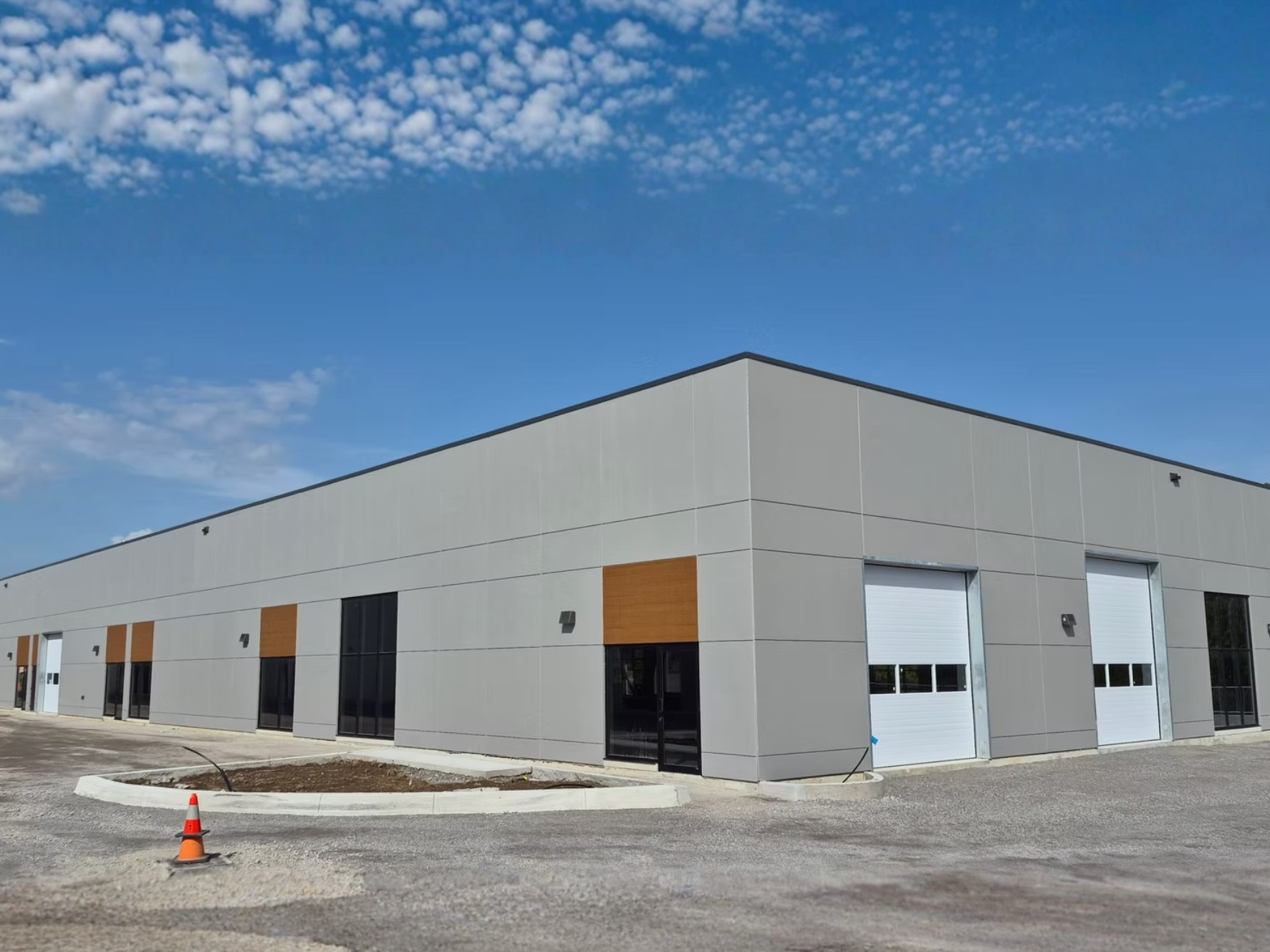 Modern commercial building with large garage doors under a blue sky with scattered clouds. The building has gray and white exterior with wooden accents and a small parking lot in the foreground.