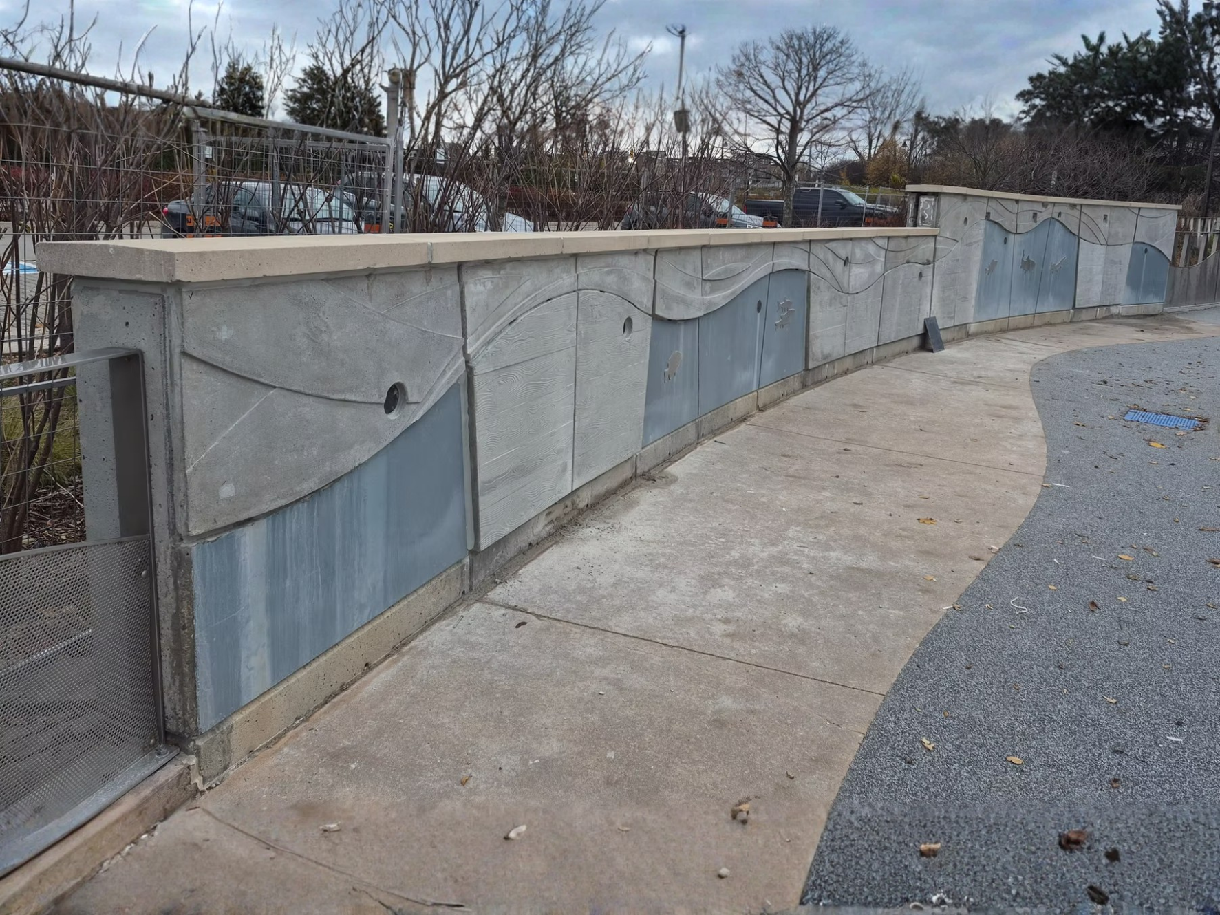 Concrete wall with decorative wave pattern and metal panels, running beside a curved asphalt and concrete sidewalk in an outdoor area. The background shows leafless trees, parked cars, and a cloudy sky.