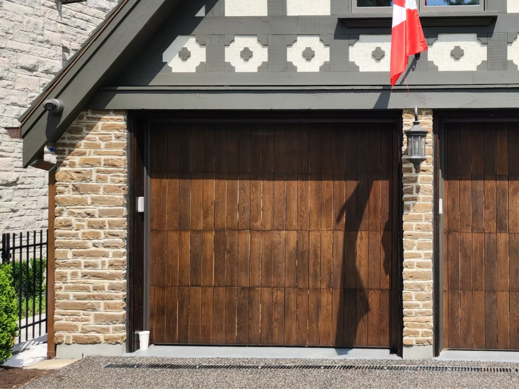 Image of a residential garage with two wooden doors, brick columns, decorative gray trim, a Canadian flag, and a lantern-style light fixture on a sunny day.