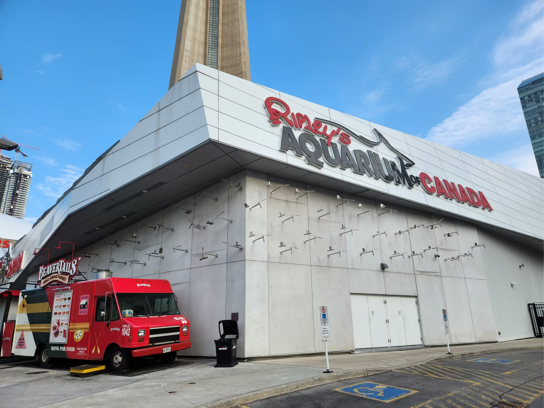 Exterior view of Ripley's Aquarium of Canada, a large modern building with a white facade and the name in bold letters. There is a red food truck parked in front, and a few parking spots marked with accessible signs. The sky is partly cloudy with some high-rise buildings visible in the background.