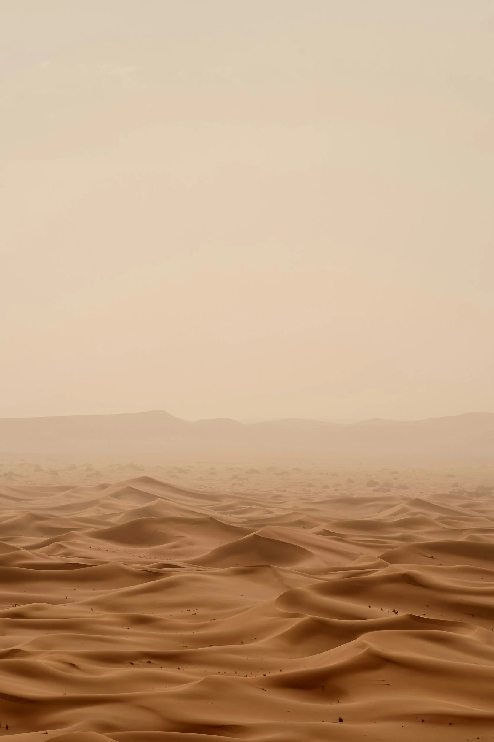 Sand dunes in a desert with mountains in the background under a hazy sky.