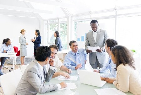 Business professionals in a modern office meeting setting, some seated around a table and others standing and conversing.