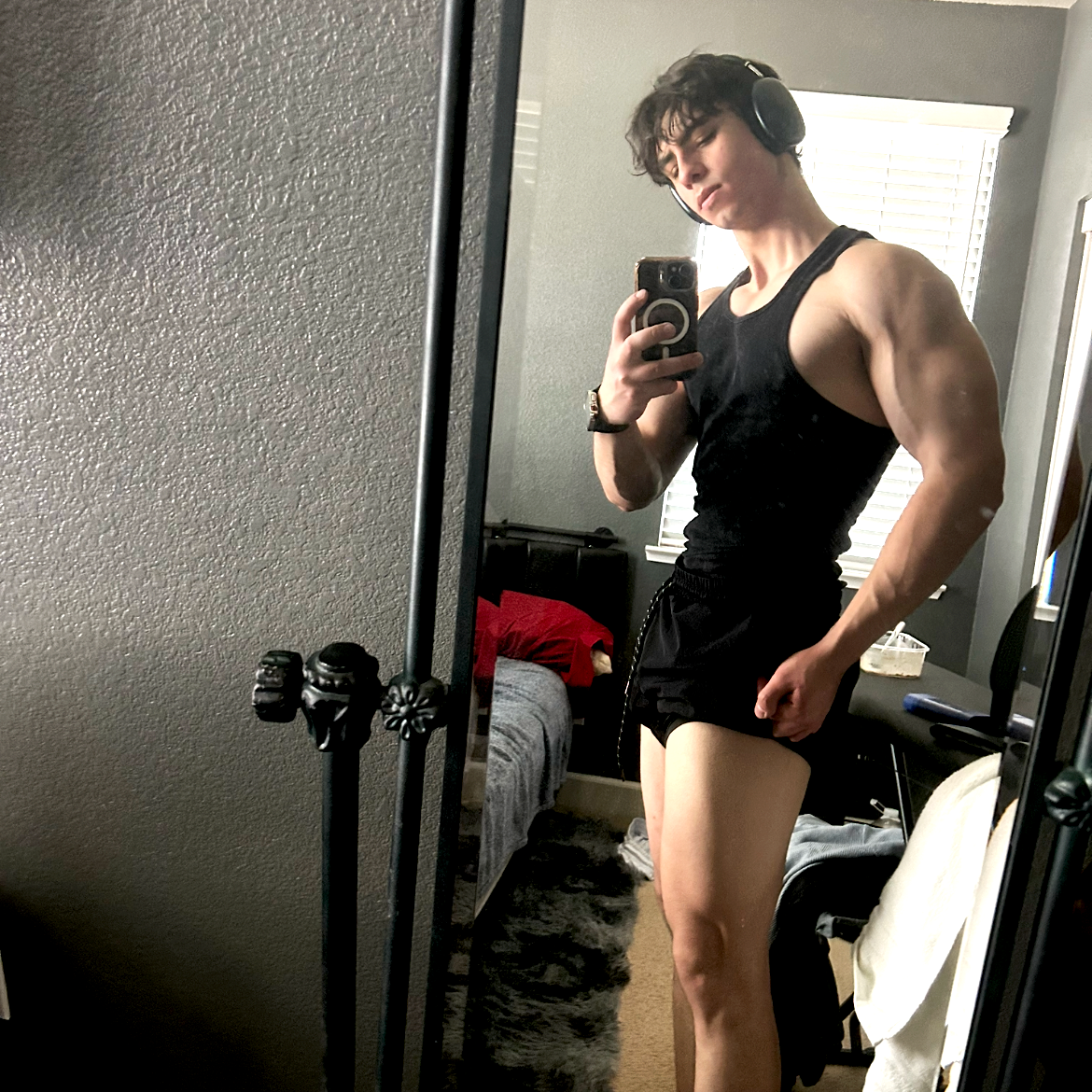 Young man taking a mirror selfie, flexing his bicep, wearing headphones, a black tank top, and black shorts in a bedroom.