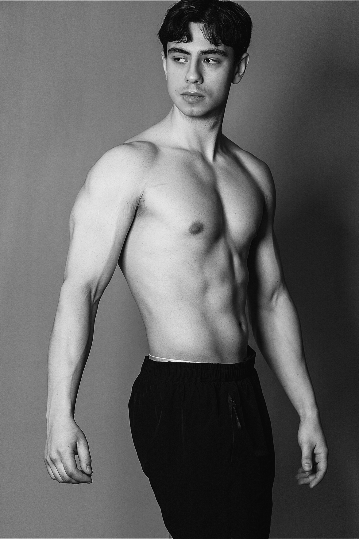 A young shirtless man with dark hair stands against a plain background, looking to his left with a neutral expression.