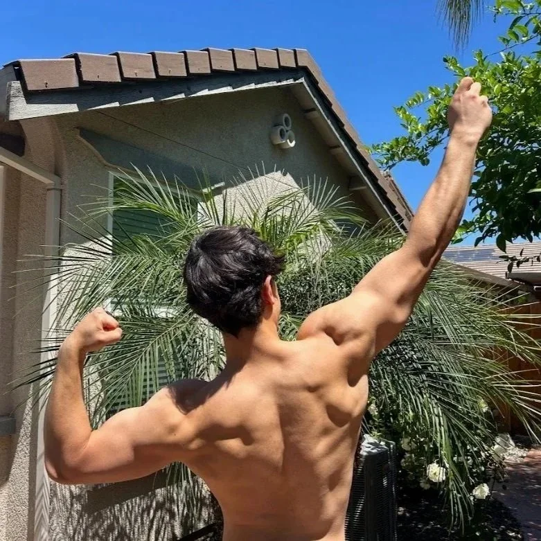 A shirtless man with dark hair flexes his back and arm muscles outdoors, standing in front of a palm tree and a house with a beige wall and a brown-tiled roof, under a clear blue sky.
