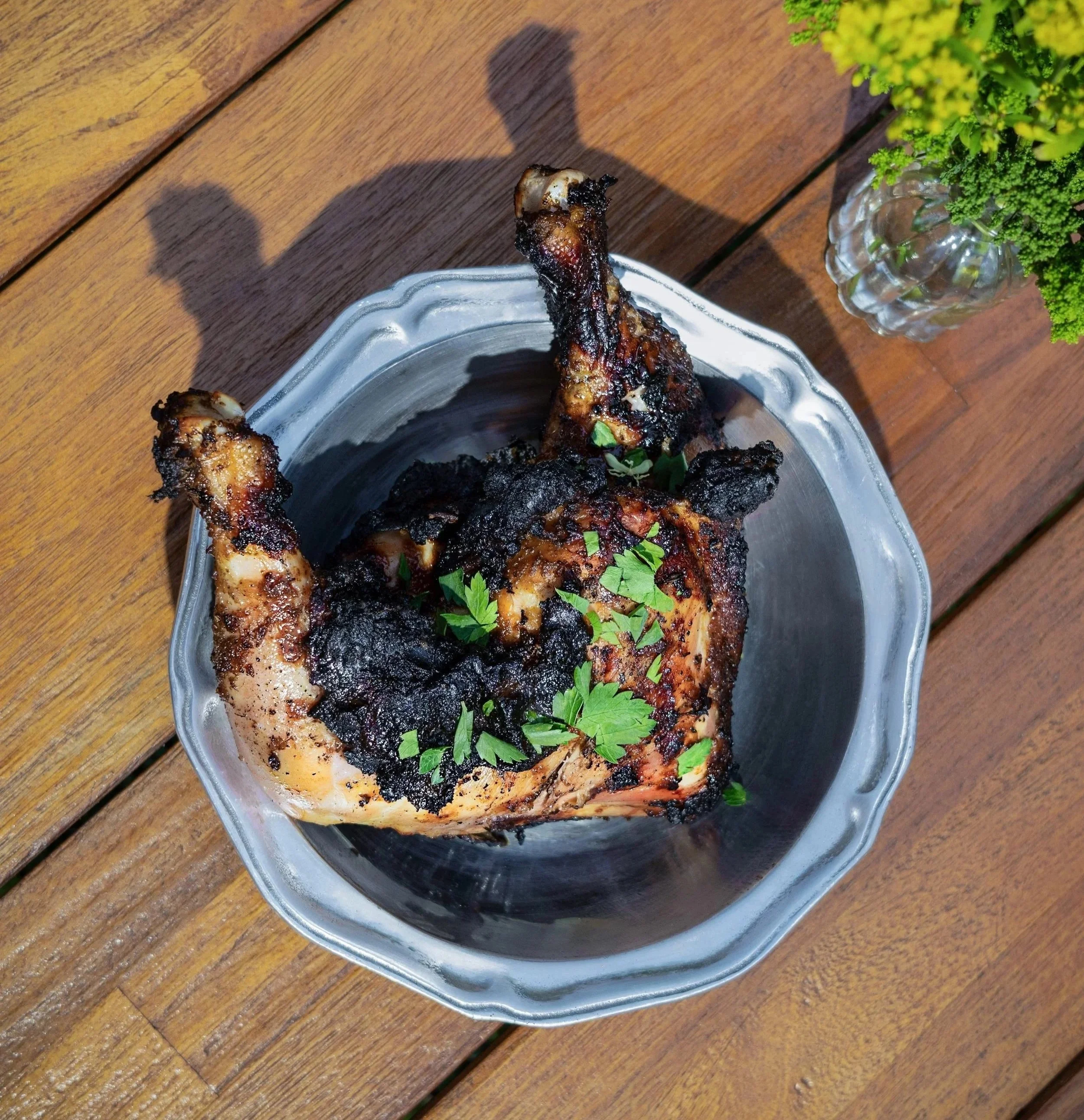 Close-up of grilled chicken drumsticks on a silver dish with parsley, on a wooden table next to a potted green plant.
