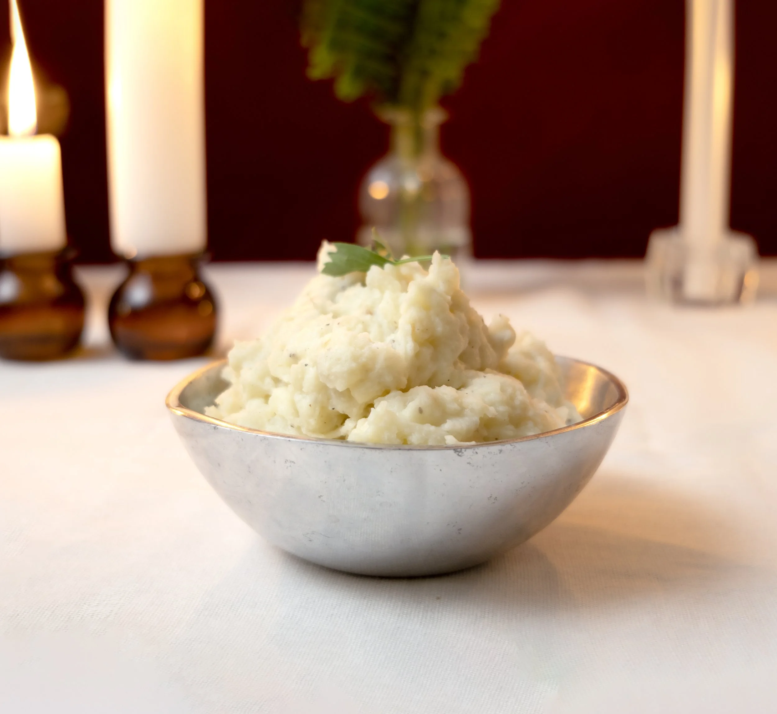 A silver bowl filled with mashed potatoes garnished with a sprig of parsley. In the background, a vase with a green leafy plant and two lit candles in holders are visible, with a red wall behind them.