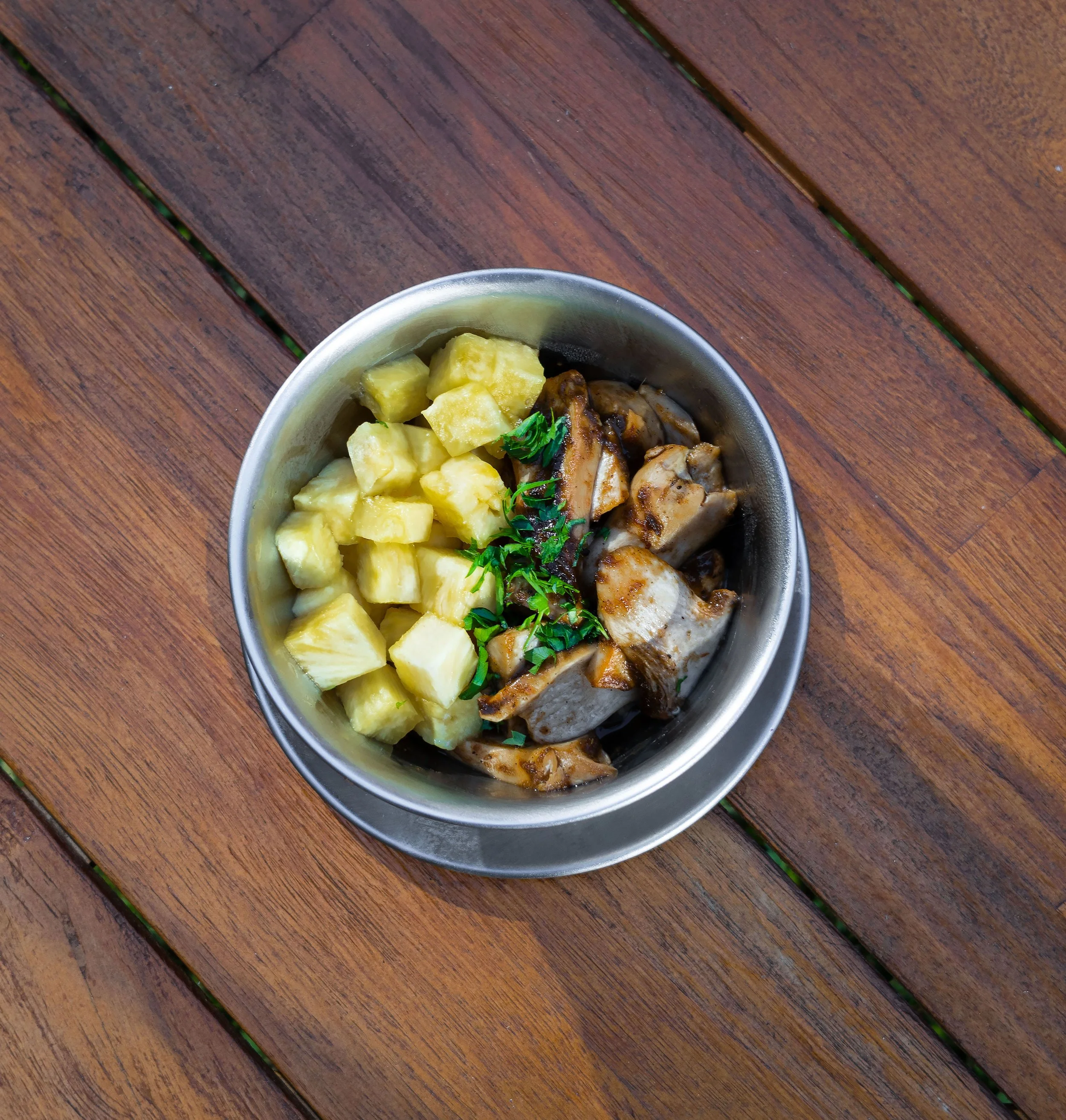 Close-up of a metal bowl containing chunks of pineapple, pieces of grilled chicken, and garnished with fresh parsley, placed on a wooden table.
