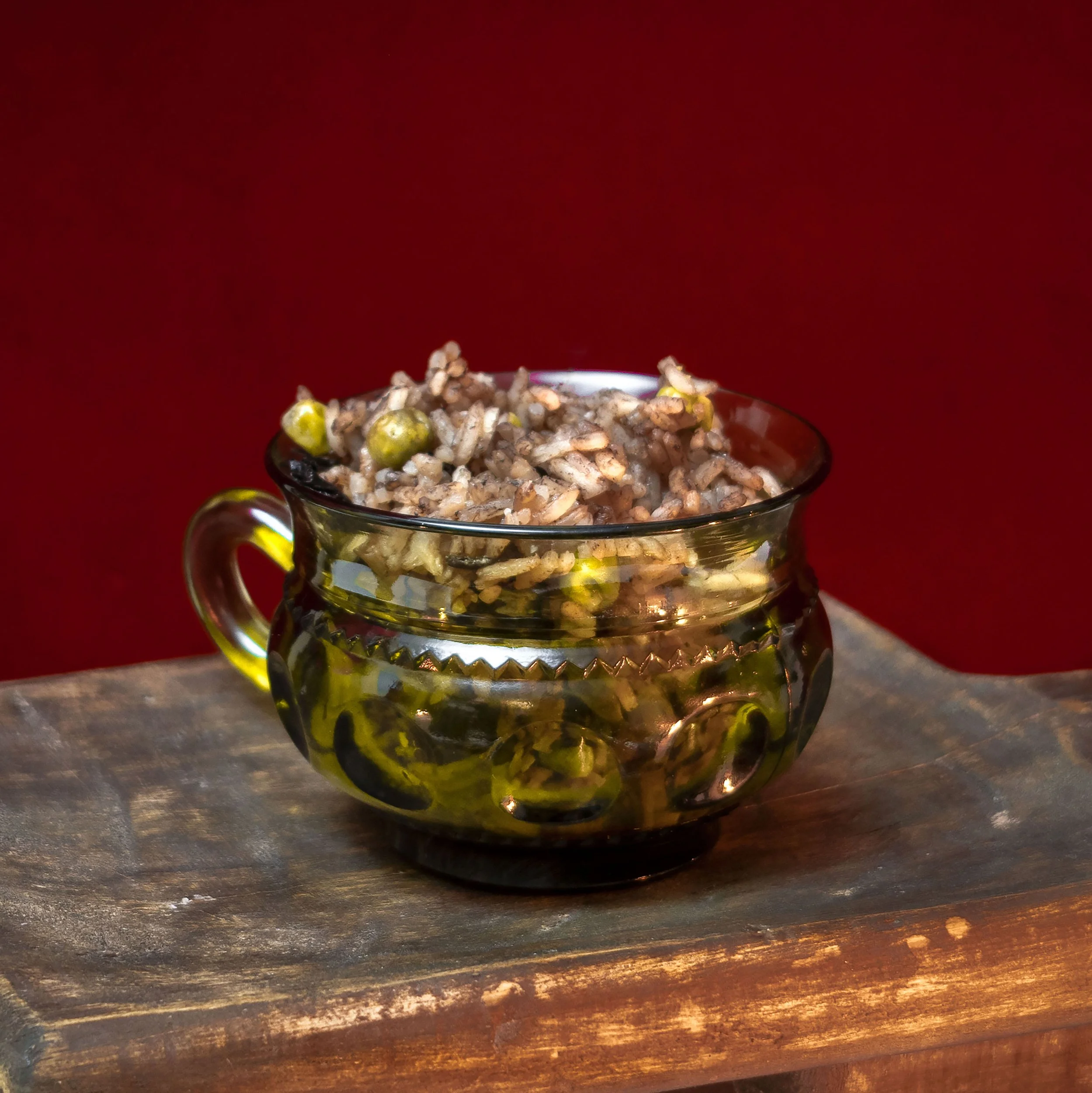 A small glass cup filled with rice and some green peas, placed on a rustic wooden surface with a red background.