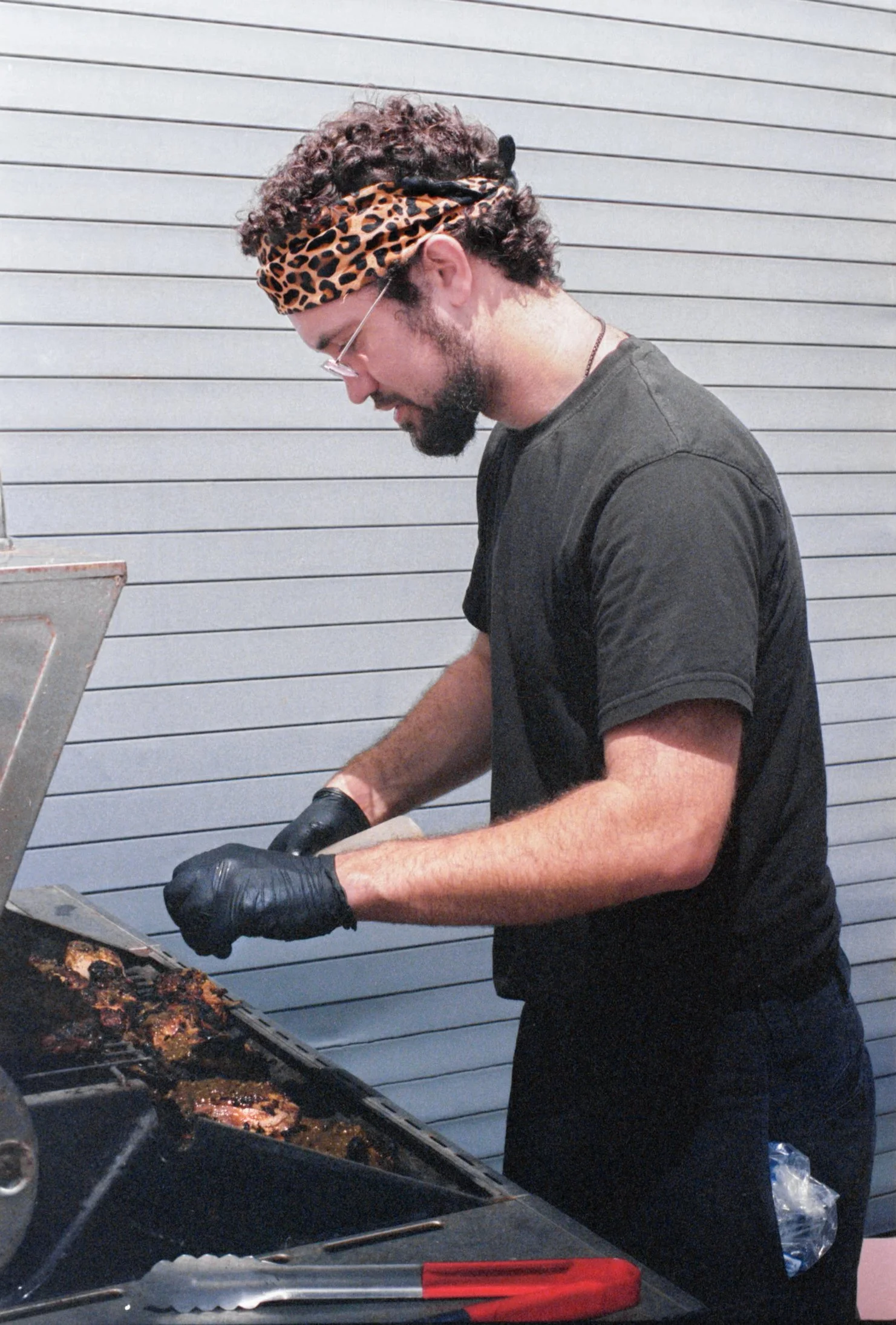 A man with curly hair, a beard, and glasses is grilling meat on a barbecue. He is wearing a leopard print headband, a black t-shirt, black gloves, and is focused on flipping or arranging the meat on the grill. The background features gray horizontal siding.