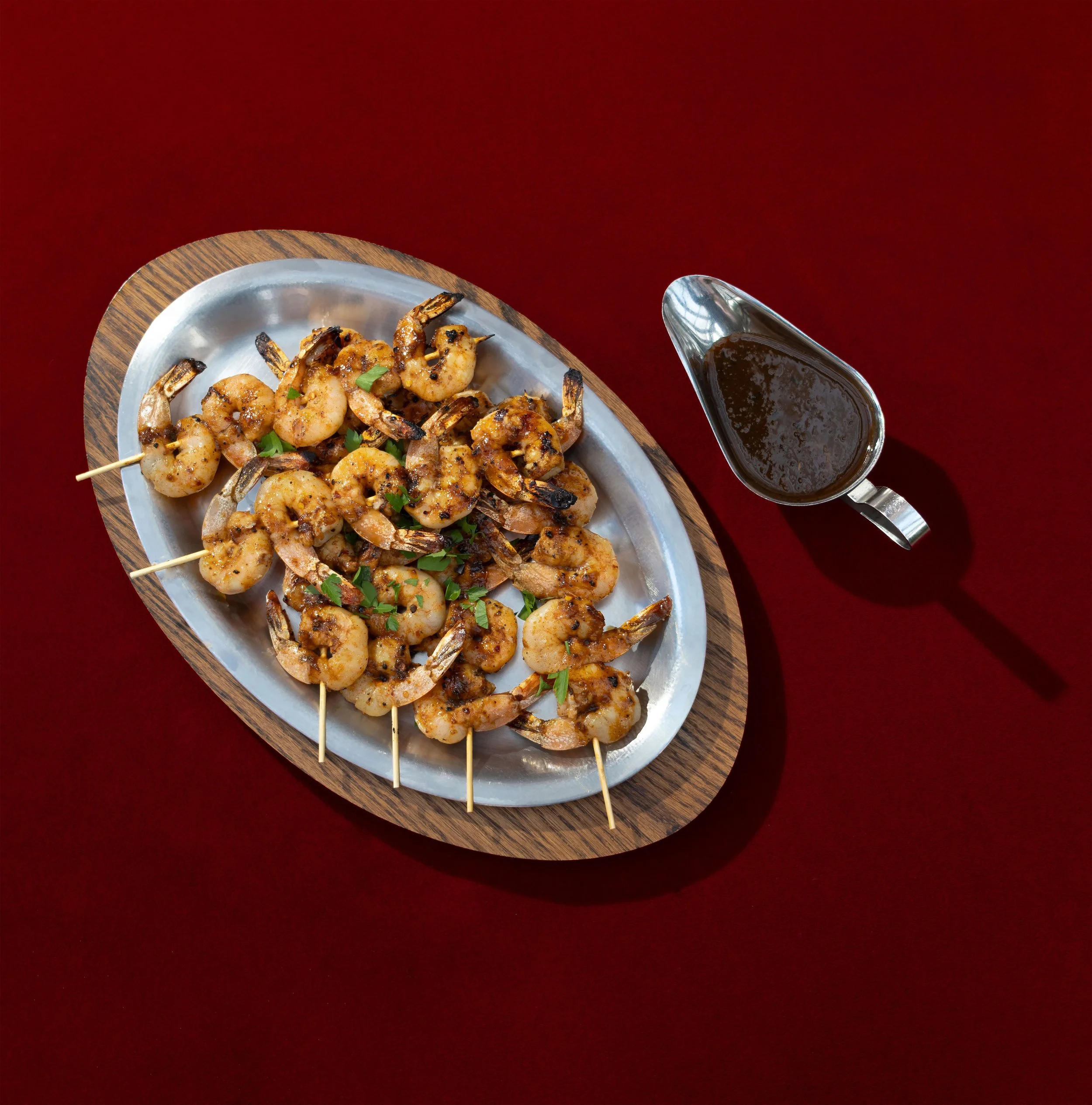 A metal tray with grilled shrimp skewers garnished with herbs, next to a pitcher of dark sauce on a wooden serving board against a red background.