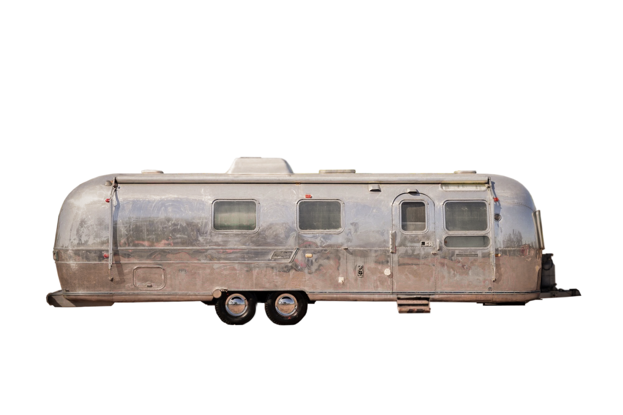 A vintage camper trailer with a weathered silver metallic exterior, three windows on the side, a small door, and dual axles with wheels, set against a black background.