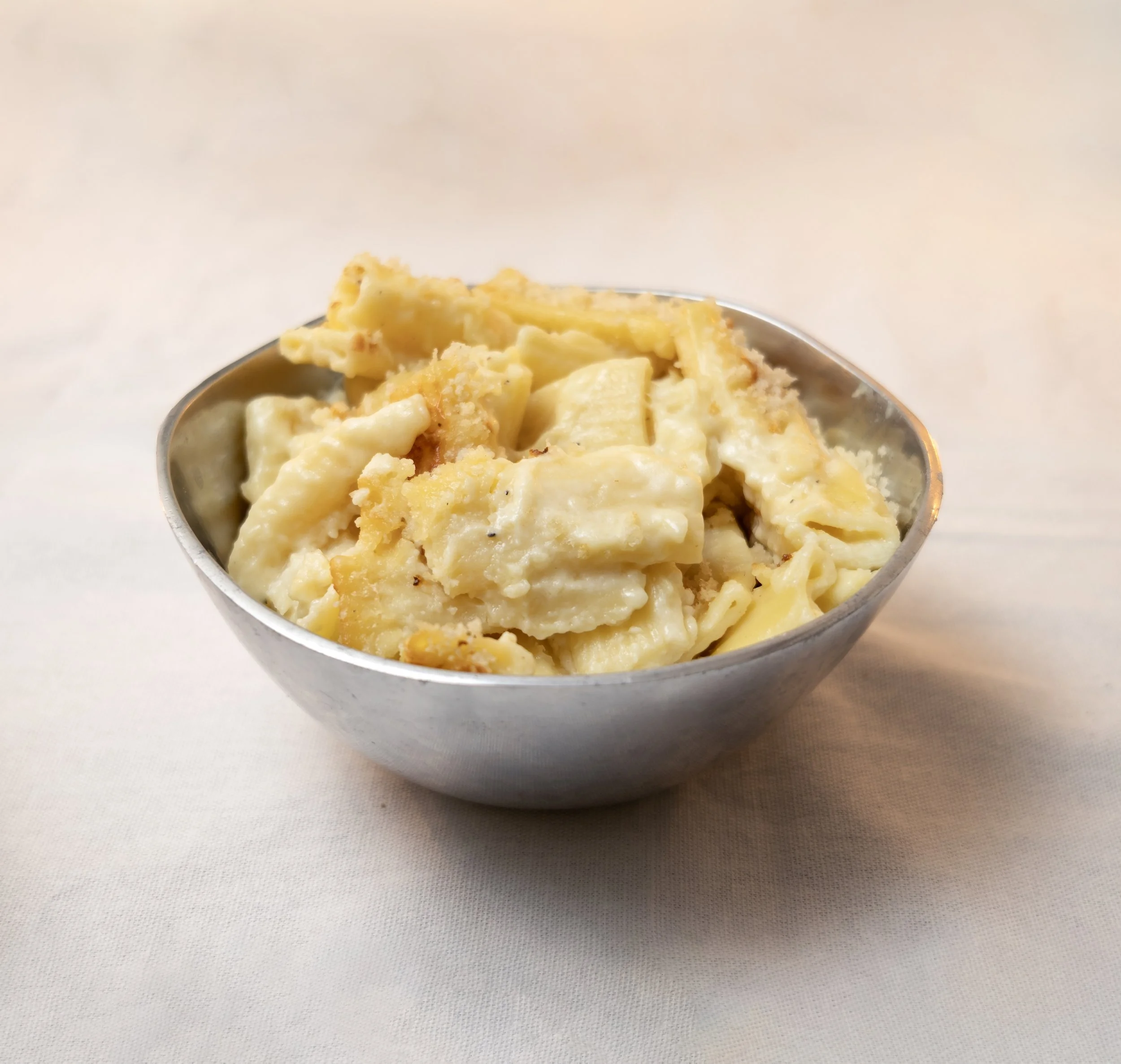 Close-up of a bowl of macaroni and cheese with a golden breadcrumb topping, on a white textured surface against a plain light background.