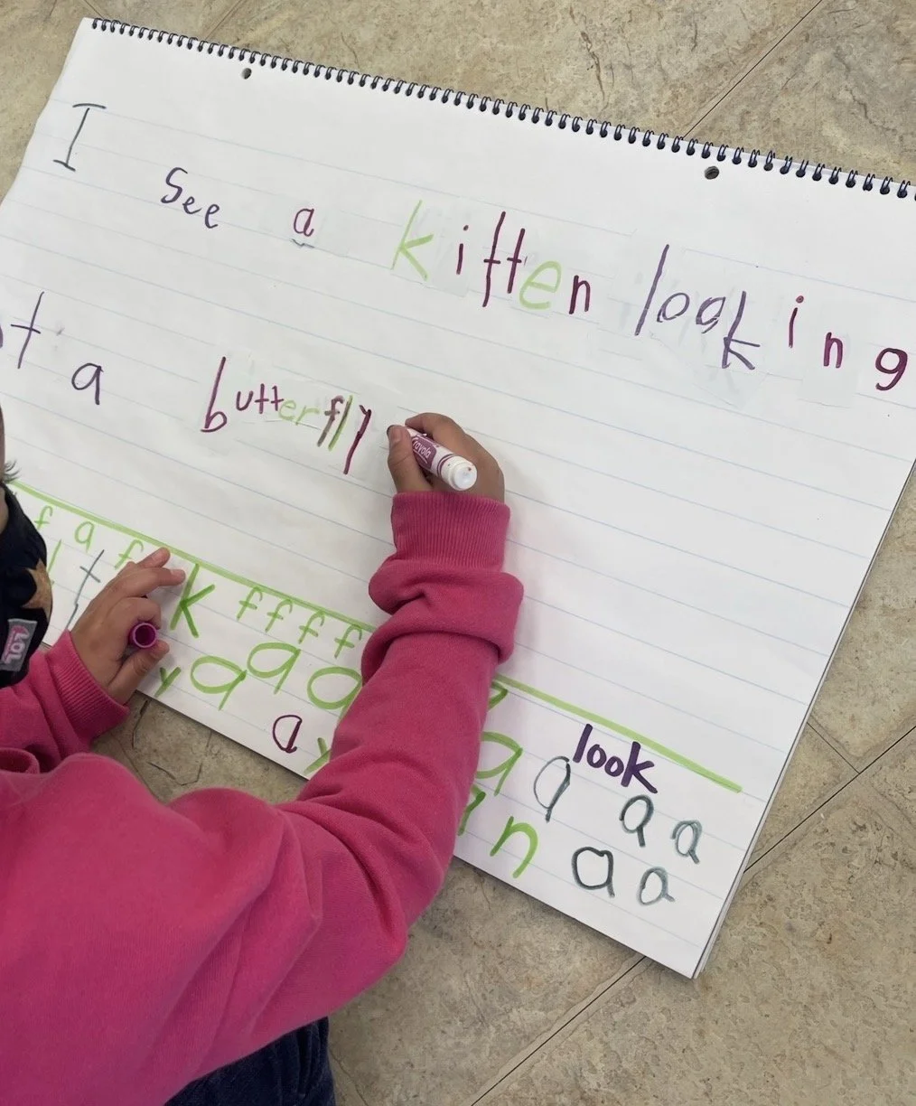 Child in a pink hoodie writing on a large lined sheet of paper with colorful marker pens, practicing spelling words like "kitten" and "butterfly" with both uppercase and lowercase letters.
